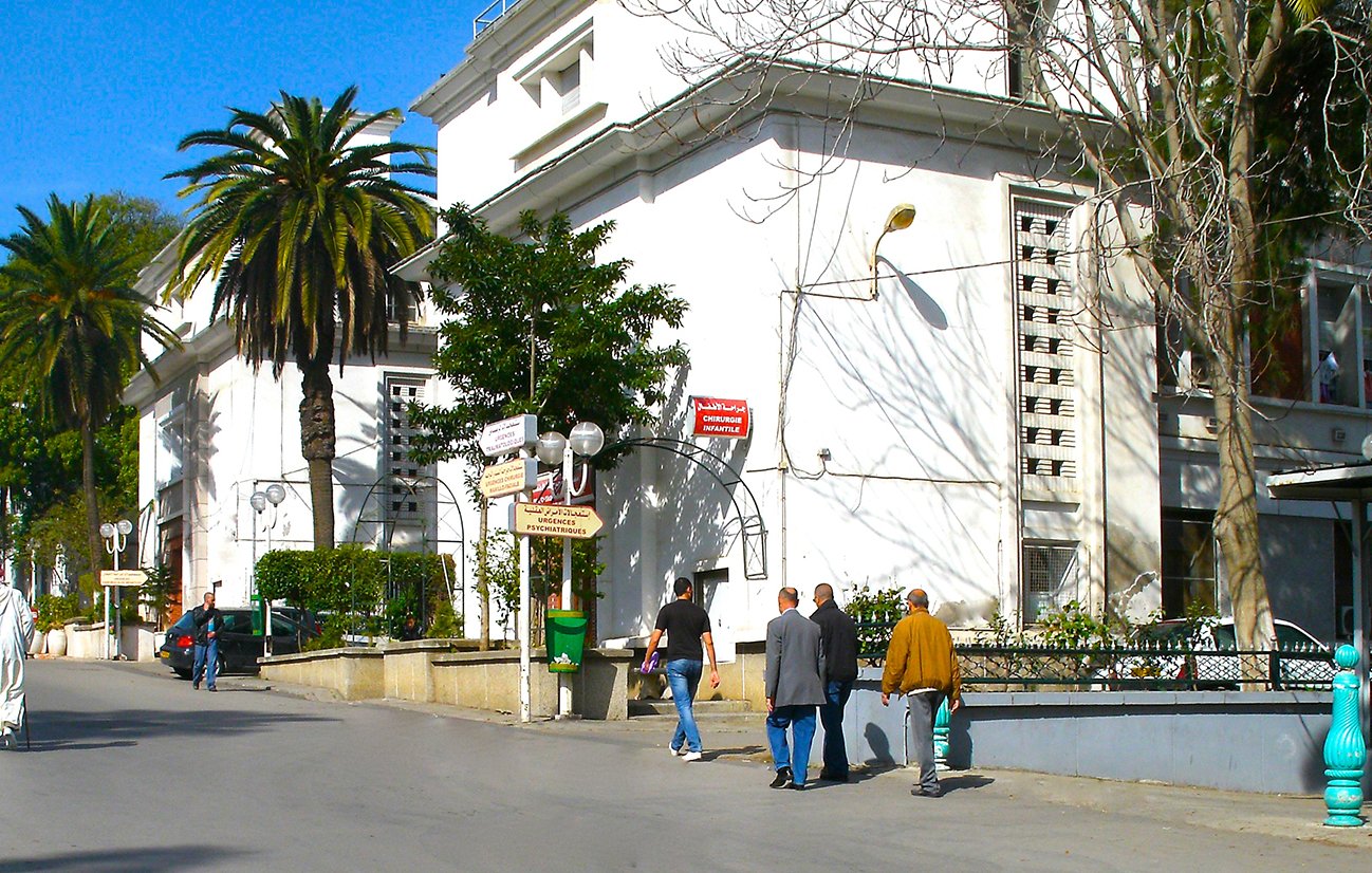 تظهر الصورة مشهداً لشارع في منطقة حضرية، حيث يمكن رؤية مباني بيضاء ذات تصميم كلاسيكي، محاطة بأشجار النخيل والمسطحات الخضراء. في الواجهة، يلاحظ وجود رجال يمشون على الرصيف، وبعض الأشخاص يرتدون ملابس تقليدية. السماء زرقاء وصافية، مما يضيف جوًا من الإشراق إلى المشهد. الجو يبدو هادئًا ومنظمًا، مع تفاصيل معمارية تبرز جمال المنطقة.