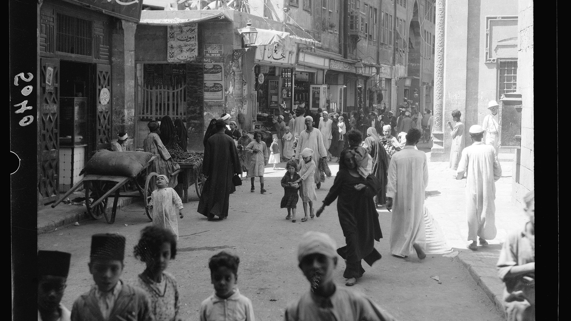 L'image montre une scène animée d'une rue dans une ville. On y voit des groupes de personnes marchant, certaines en conversation. Des enfants jouent au milieu des passants. L'architecture autour d'eux suggère un paysage urbain traditionnel, avec des bâtiments ornés et des boutiques. Il y a aussi une charrette et des femmes et hommes vêtus de vêtements traditionnels. L'ambiance générale semble dynamique et vivante, reflétant la culture locale.