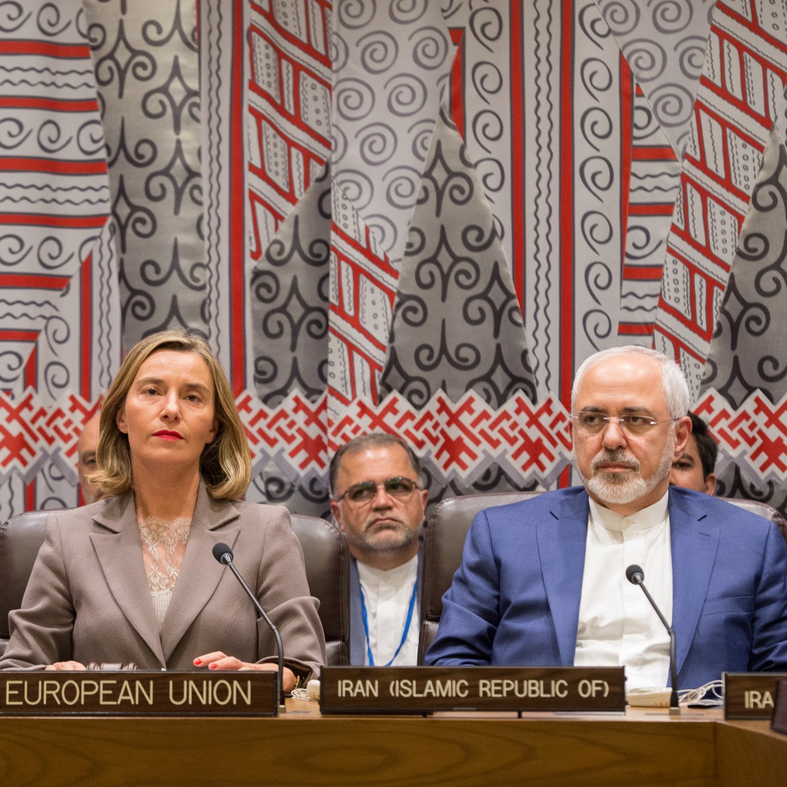 L'image montre une réunion internationale avec plusieurs personnes assises à une table. On peut voir des panneaux identifiant les différents participants, tels que l'Union européenne et l'Iran. Trois figures centrales semblent être des représentants importants, avec des expressions sérieuses. En arrière-plan, il y a des individus qui écoutent attentivement. Le décor est riche et coloré, avec des motifs géométriques sur le mur derrière eux, suggérant un cadre formel pour des discussions diplomatiques.
