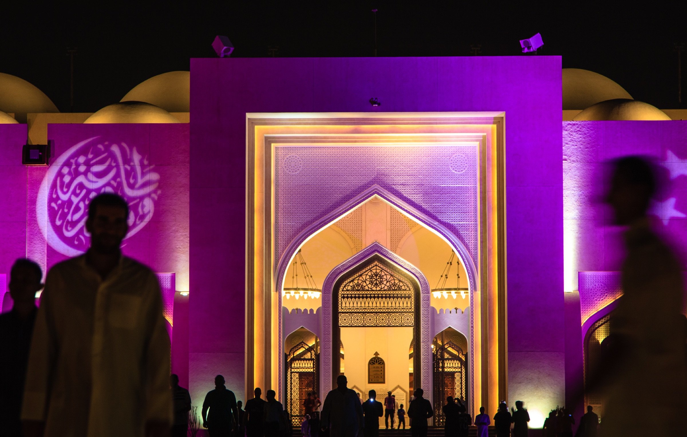 L'image montre une grande structure architecturale éclairée de manière colorée, principalement en violet. On peut voir un portail monumental avec des motifs décoratifs, typiques de l'architecture islamique. À l'avant, des silhouettes de personnes marchent, créant une atmosphère vivante et dynamique. Au fond, des éléments de design comme des arcs et des motifs sont visibles, ajoutant une touche d'élégance à l'ensemble de la scène. L'éclairage nocturne met en valeur les détails architecturaux et crée une ambiance fascinante.