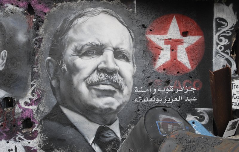 L'image montre un graffiti sur un mur, représentant un portrait en noir et blanc d'un homme avec une moustache et un regard déterminé. À côté du portrait, on peut voir une étoile rouge avec un symbole. Des inscriptions en arabe sont également visibles, accompagnées de motifs colorés sur le mur. L'ensemble donne une impression d'engagement politique, avec une ambiance urbaine et artistique.