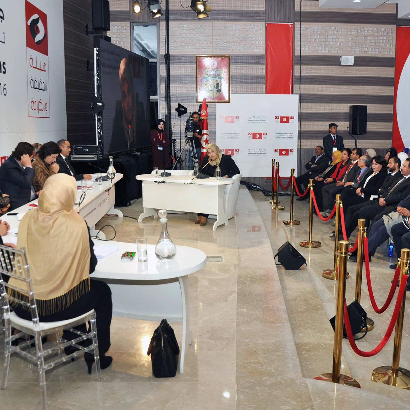L'image montre un panel de discussion ou une session d'audition, avec plusieurs personnes assises face à une table à l'avant. Au fond, il y a une bannière affichant des informations sur l'événement, datée des 16 et 17 décembre 2016. Les participants semblent engagés dans la discussion, avec une variété de personnes, y compris des femmes portant des foulards. La scène est bien organisée, avec des cordons de sécurité délimitant l'espace où se trouvent le public et les intervenants.