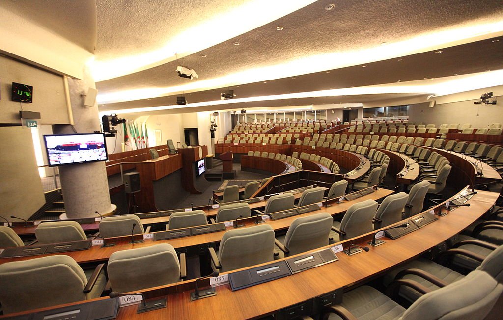 L'image montre une salle de réunion ou un hémicycle, probablement utilisée pour des sessions parlementaires ou des assemblées. On peut voir des rangées de sièges confortables disposés en arc de cercle, avec des tables en bois devant chaque siège. Des écrans et des équipements audiovisuels sont visibles, indiquant que la salle est équipée pour des présentations ou des débats. Les murs sont décorés de manière sobre et moderne, créant une ambiance institutionnelle.