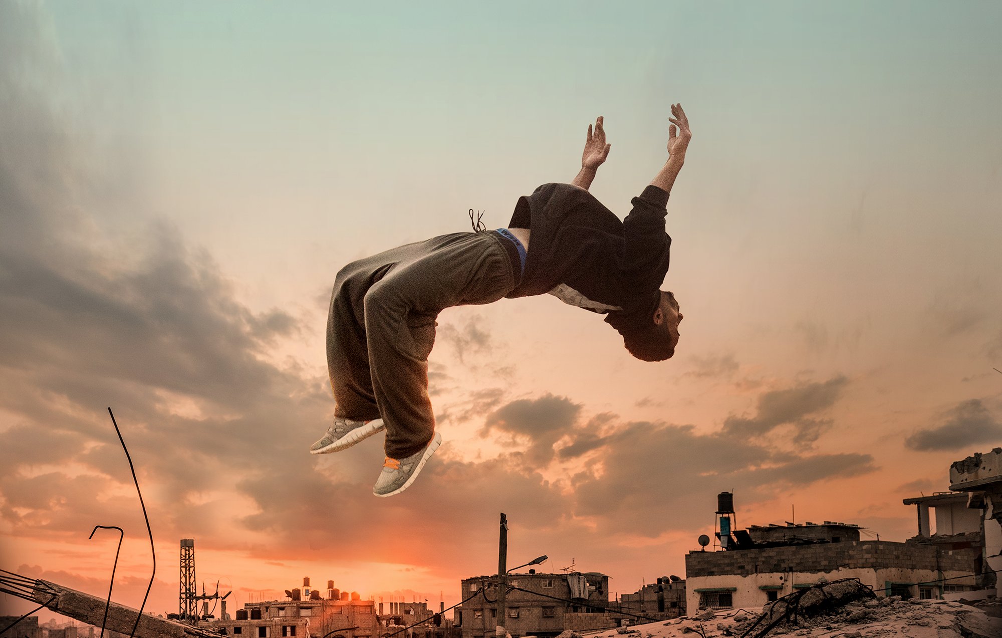 L'immagine mostra un giovane che sembra fluttuare o saltare sopra un paesaggio urbano distrutto, con scarti di costruzione e rovine. Sullo sfondo, si possono vedere nuvole e un cielo al tramonto, che creano un'atmosfera drammatica. La silhouette del ragazzo è evidenziata dalla luce, evidenziando un senso di libertà e movimento in un contesto desolato.
