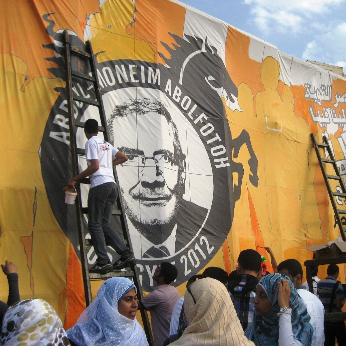 L'image montre un groupe de personnes en train de peindre une grande bannière ou une affiche. Ils sont sur des échelles et travaillent à mettre en place un portrait ou un dessin qui semble représenter une figure importante, peut-être liée à un événement politique. Le fond est coloré, principalement en orange, et il y a des inscriptions en arabe. Les femmes au premier plan portent des foulards, et l'atmosphère semble dynamique, avec des gens engagés dans le travail collectif.