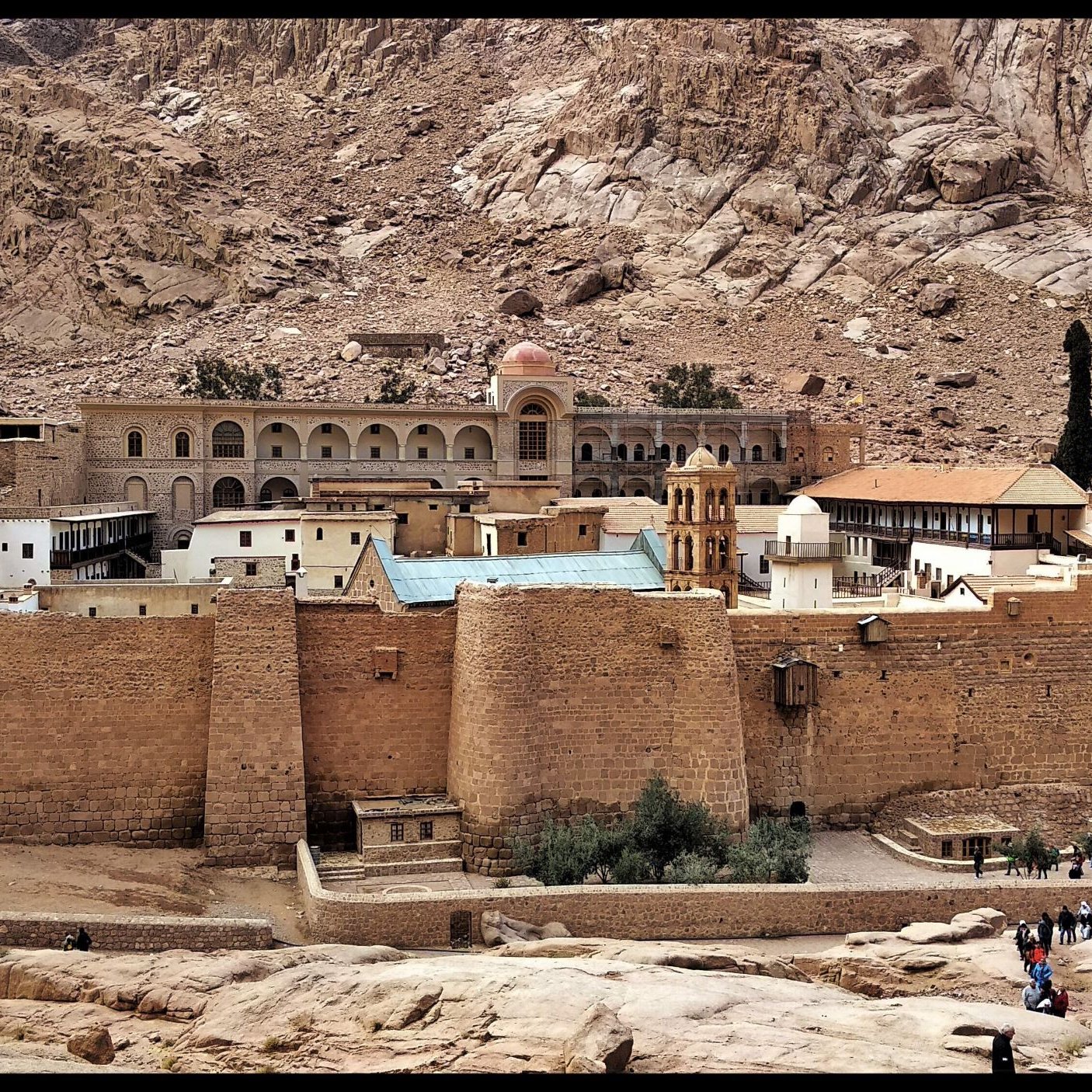 L'image montre un monastère situé dans une région montagneuse aride. Le bâtiment, de style architectural ancien, est construit en pierre et possède des murs épais. On peut voir une grande cour intérieure ainsi que des arbres qui ajoutent une touche de verdure au paysage. À l'arrière-plan, on distingue des formations rocheuses spectaculaires, soulignant l'isolement et la beauté naturelle des lieux. Des visiteurs se promènent autour du monastère, ce qui témoigne de son attrait touristique.
