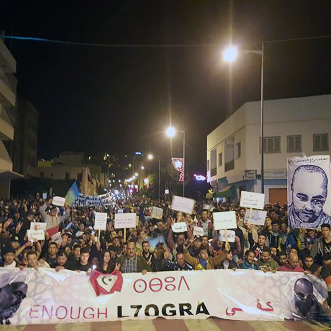 The image depicts a large crowd gathered in a street at night, participating in a protest or demonstration. Many individuals hold signs, and a prominent banner at the front reads "ENOUGH L7OGRA." There is also a large portrait of a person displayed, likely related to the cause of the protest. The atmosphere appears charged, with people expressing their views collectively in a public space. Streetlights illuminate the scene, highlighting the participants and their messages.