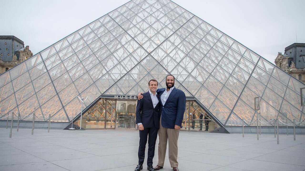 L'image montre deux hommes posant ensemble devant le Musée du Louvre, qui est célèbre pour sa pyramide en verre. Le bâtiment est moderne et impressionnant, contrastant avec l'architecture classique des ailes du musée. Les hommes sourient et portent des costumes formels, se tenant côte à côte, tandis que le ciel est légèrement nuageux, ajoutant une atmosphère calme à la scène.