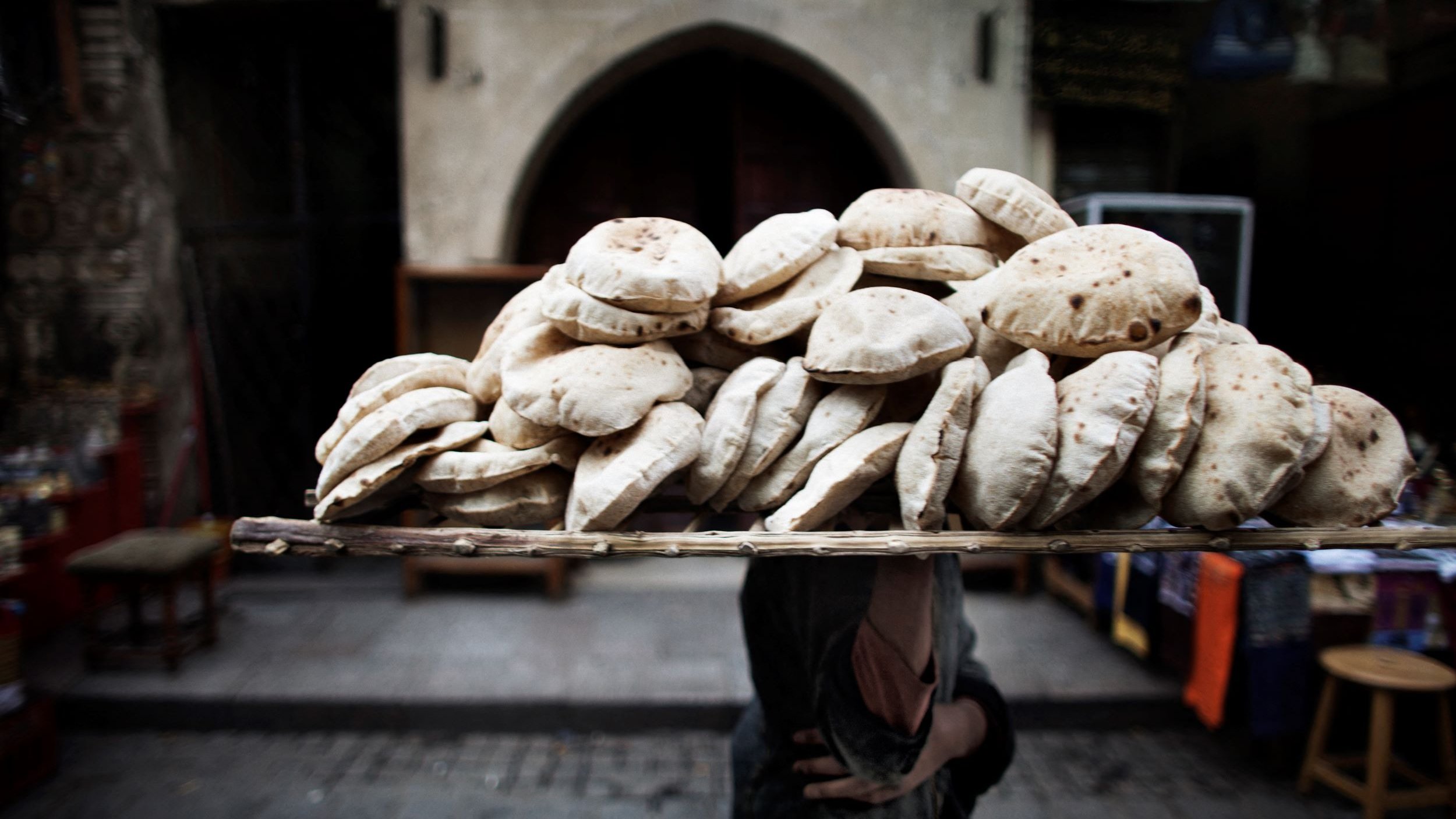 تظهر في الصورة مجموعة كبيرة من الخبز العربي، المعروف باسم "الخبز المسطح" أو "العربات". يحمِل شخصٌ ما سلة محملة بهذه الأرغفة على رأسه، مما يعكس طريقة تقليدية لنقل الطعام في بعض الأسواق. الخلفية تحتوي على معالم معمارية قديمة، مما يوحي بأن الصورة تم التقاطها في منطقة تاريخية أو سوق شعبي. الجو العام يوحي بالنشاط والحياة اليومية في هذا المكان.