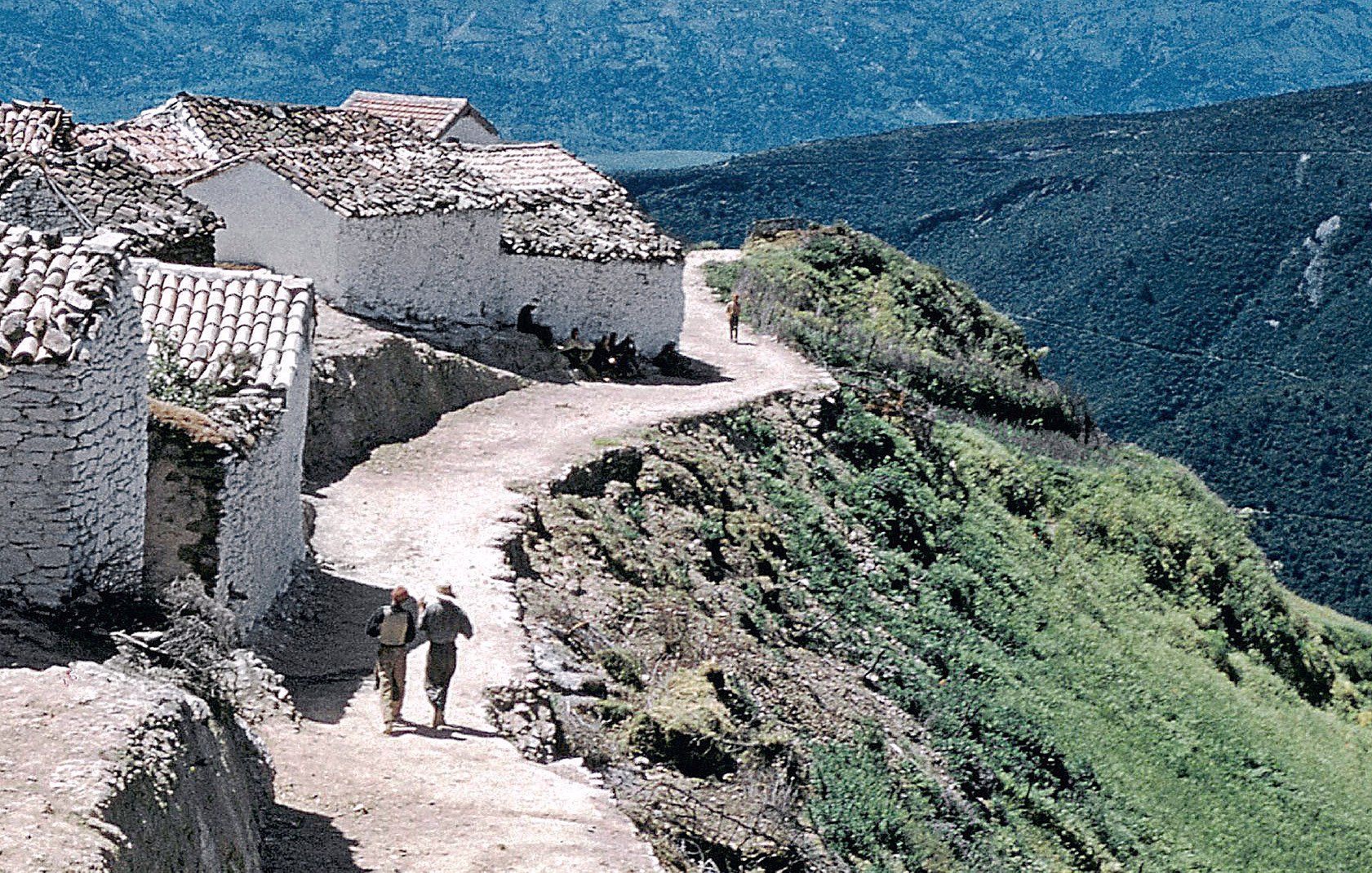 L'image montre un paysage rural avec un sentier serpentant le long d'une colline. De chaque côté du chemin, on peut voir des maisons blanches avec des toits en tuiles. Deux personnes marchent sur le sentier, profitant de la vue. En arrière-plan, des montagnes verdoyantes s'élèvent, créant une atmosphère paisible et naturelle. Le ciel est clair et contribue à l'ambiance sereine de la scène.