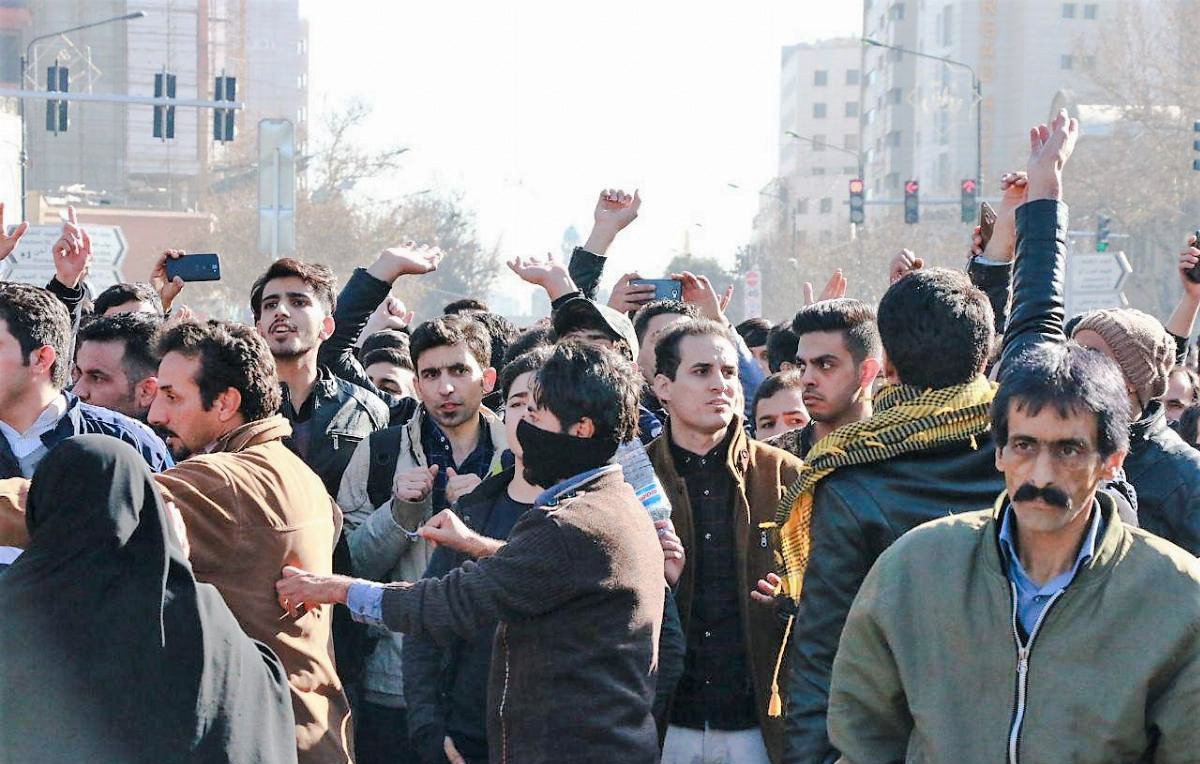 تظهر في الصورة مجموعة من الناس يتجمعون في مكان مفتوح، يبدو أنهم يشاركون في تظاهرة أو احتجاج. هناك مجموعة متنوعة من الأشخاص في الصورة، منهم رجال ونساء، وبعضهم يظهر بوجوه مكشوفة بينما البعض الآخر يلبس أغطية على وجهه. خلفهم يمكن رؤية لافتات وإشارات، مما يشير إلى أنهم يعبرون عن رأي أو موقف معين. الجو العام يوحي بالتحمس والجدل، حيث يبدو أن البعض يرفعون هواتفهم لتوثيق الحدث.