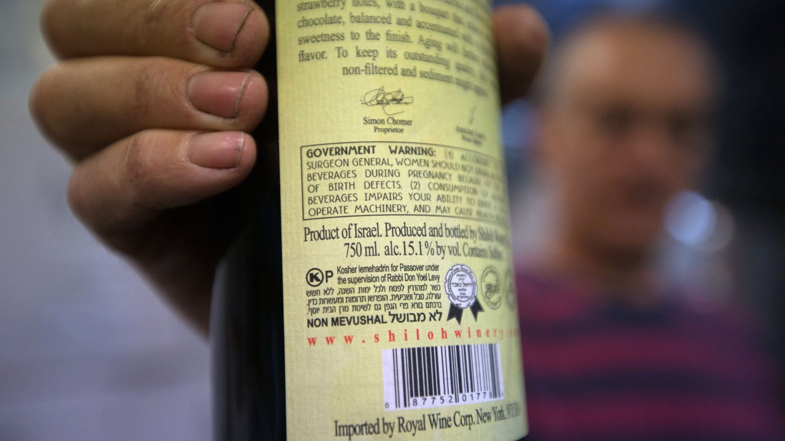 L'image montre une personne tenant une bouteille de vin, avec l'étiquette clairement visible. L'étiquette comporte des informations détaillées sur le produit, y compris des avertissements et des mentions sur la provenance, indiquant qu'il s'agit d'un vin produit en Israël. La bouteille est de 750 ml et a un taux d'alcool de 15,1 % vol. En arrière-plan, on peut deviner une autre personne floue, mais les détails ne sont pas très clairs. Les mains de la personne tenant la bouteille semblent légèrement sales, ce qui pourrait suggérer un environnement de travail ou une activité en plein air.