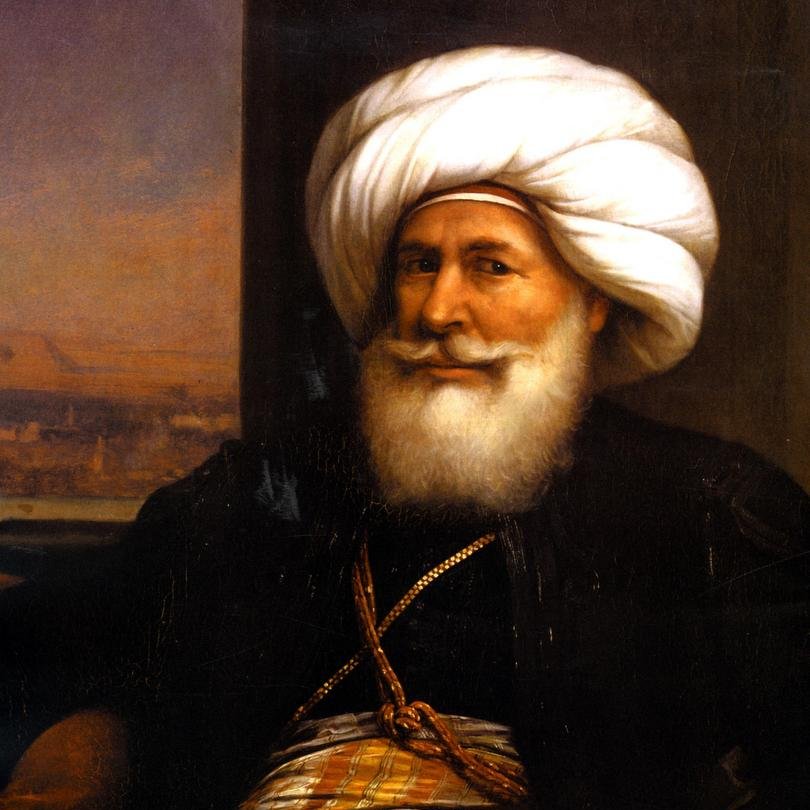 تظهر الصورة رجلاً مسنًّا يرتدي عمامة كبيرة ومجموعة من الملابس التقليدية. يتميز بلحيته البيضاء الكثيفة وابتسامة ودودة. يحمل سيفًا في يده، مما يوحي بمكانته العالية أو دوره القيادي. الخلفية تحتوي على مشهد يبدو كأنه منظر طبيعي لمدينة أو تجمع حضري. تعكس تفاصيل ملابسه وزينه التراث الثقافي لمنطقته.