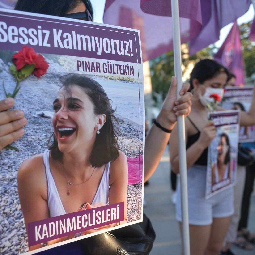 تظهر الصورة مجموعة من النساء أثناء تظاهرهن. تحمل إحداهن لافتة مكتوب عليها "Sessiz Kalmıyoruz!" (لا نصمت!) وصورة لامرأة تُدعى "Pınar Gültekin". كما تحمل إحدى المتظاهرات زهرة حمراء، مما يشير إلى أن المظاهرة تتعلق بقضية اجتماعية أو حقوق النساء. يُظهر المشهد روح التضامن والمطالبة بالعدالة.