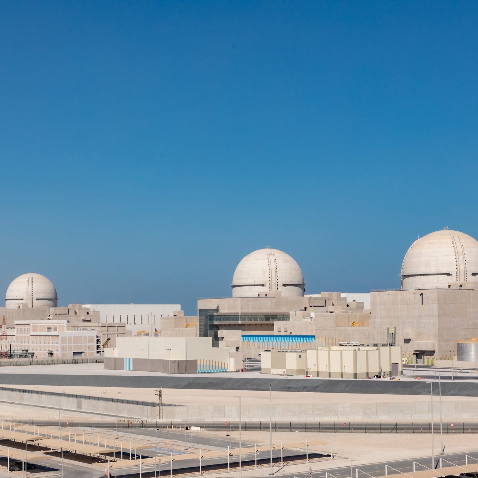 The image shows a large industrial facility, likely a nuclear power plant, with several prominent dome-shaped structures. The plant features a modern design with various buildings and infrastructure surrounding it, set against a clear blue sky. The setting appears to be an arid or semi-arid environment, with minimal vegetation. There are parking areas and some infrastructure elements visible in the foreground, indicating it is a well-developed site.