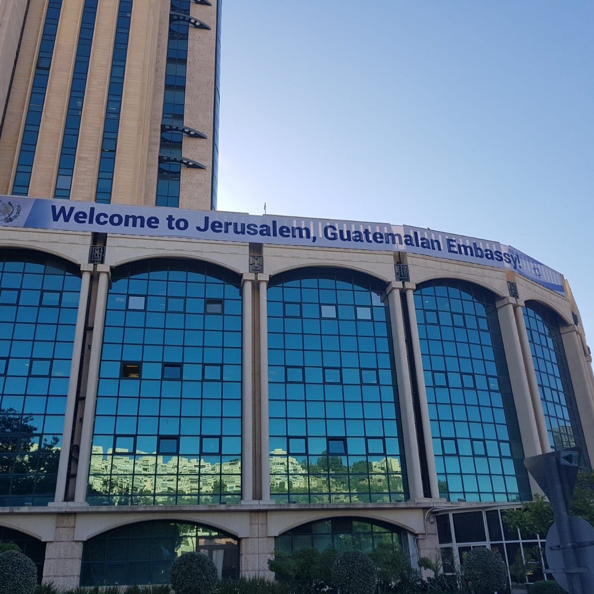 L'immagine mostra un edificio con una grande facciata di vetro riflettente. Sulla parte superiore dell'edificio è affisso uno striscione che dice "Welcome to Jerusalem, Guatemalan Embassy". Il contesto suggerisce che si tratta della sede dell'ambasciata del Guatemala a Gerusalemme. Si può notare il cielo blu sullo sfondo e alcune piante o alberi nella parte inferiore dell'immagine.