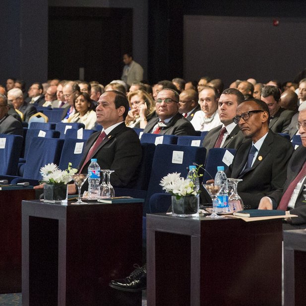 L'image montre une assemblée de personnes assises dans une salle de conférence. Les participants semblent attentifs et engagés, avec des tables devant eux, ornées de petites fleurs et de bouteilles d'eau. On peut voir plusieurs hommes en costume, ainsi que des femmes portant des vêtements formels. L'environnement est élégant, avec des chaises bleues et une disposition ordonnée. Les visages des participants expriment une concentration sur l'événement en cours.
