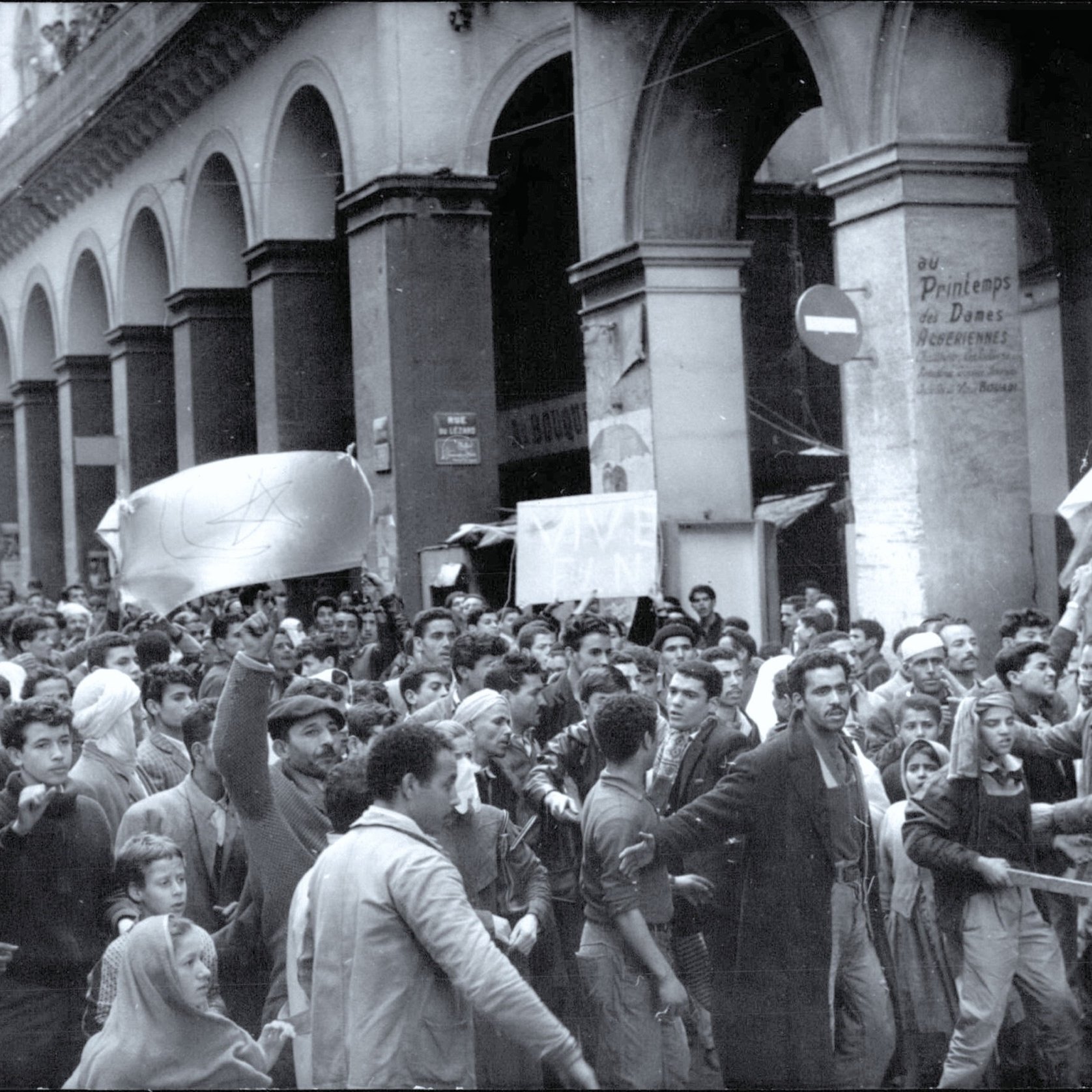 الصورة تظهر تجمعًا حاشدًا لأشخاص يسيرون في أحد الشوارع. الشارع يبدو مزدحمًا بالمتظاهرين، حيث يحمل البعض لافتات وأعلام. يبدو أنهم يعبرون عن مطالب أو احتجاجات. العمارة في الخلفية تعكس طابعًا تاريخيًا وتساهم في خلق جو من الزمان والمكان الذي تتواجد فيه هذه الفعالية. تعبيرات الوجوه تُظهر الحماس والعزيمة بين المشاركين.
