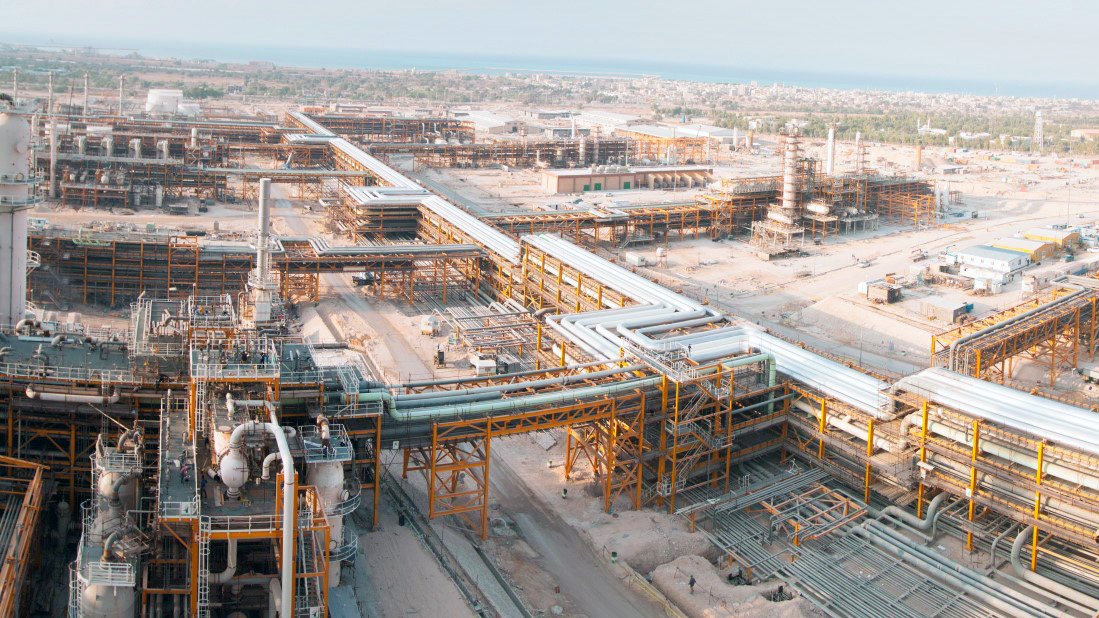 الصورة تُظهر مشهدًا واسعًا لمجمع صناعي، حيث تظهر مرافق ومعدات ضخمة. يمكن رؤية أنابيب كبيرة تمتد عبر الموقع، بالإضافة إلى أفران ومنشآت أخرى تشير إلى نشاط صناعي مكثف. الخلفية تتضمن المساحات المفتوحة والمباني التي تدل على طبيعة العمل في هذا المكان. السماء تبدو صافية، مما يضفي طابعًا مشمسًا على الصورة.
