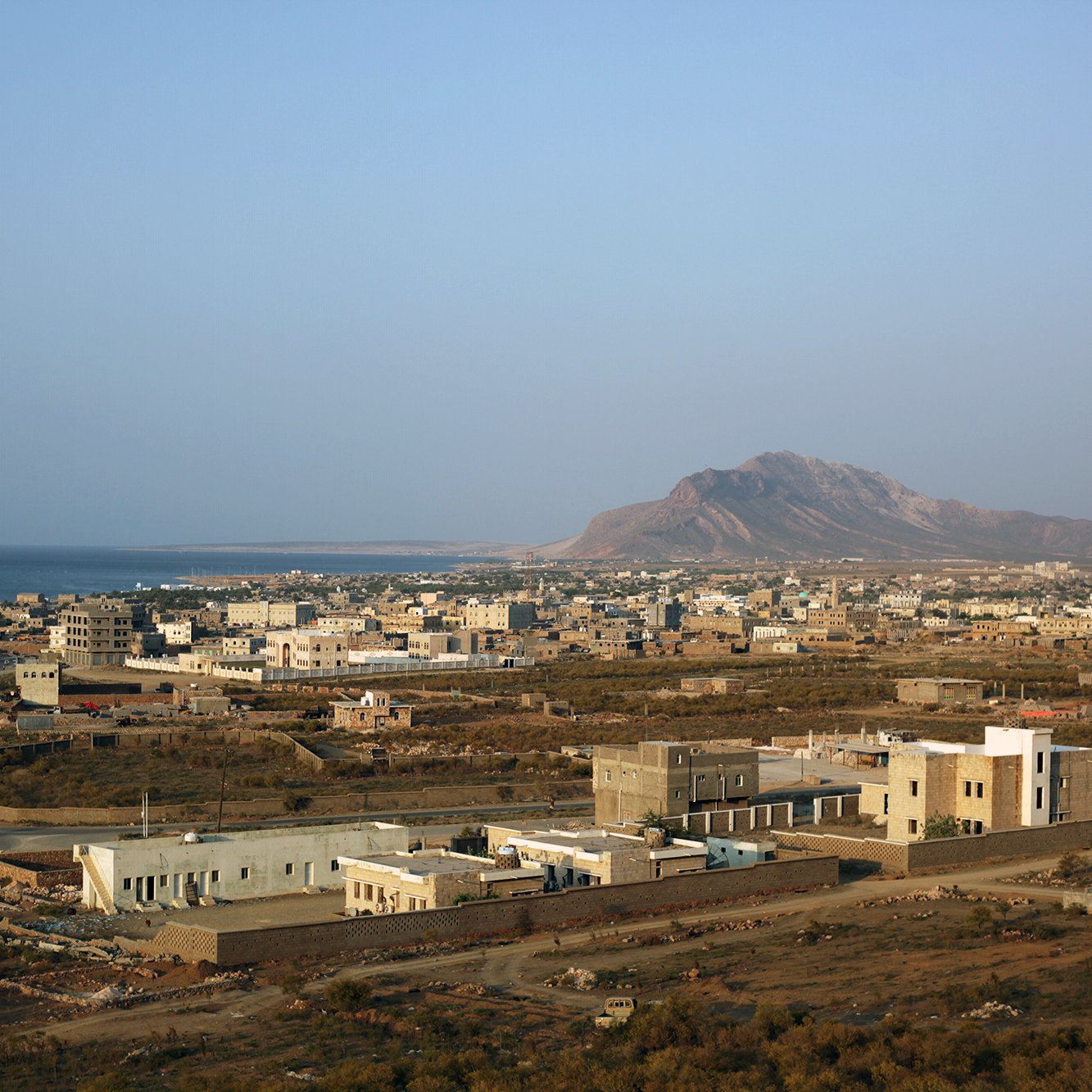 تظهر الصورة منظرًا خلابًا لبلدة ساحلية تطل على البحر. يُظهر المشهد منازل مبنية بشكل متفرق، بعضها حديث والآخر تقليدي، مع شوارع تمتد بين المباني. في الخلفية، يمكن رؤية سلسلة جبال، بينما يمتد البحر الفسيح على الجهة اليسرى. السماء صافية وتبدو أشعة الشمس مشرقة، مما يضفي شعورًا بالهدوء والجمال الطبيعي على المكان.