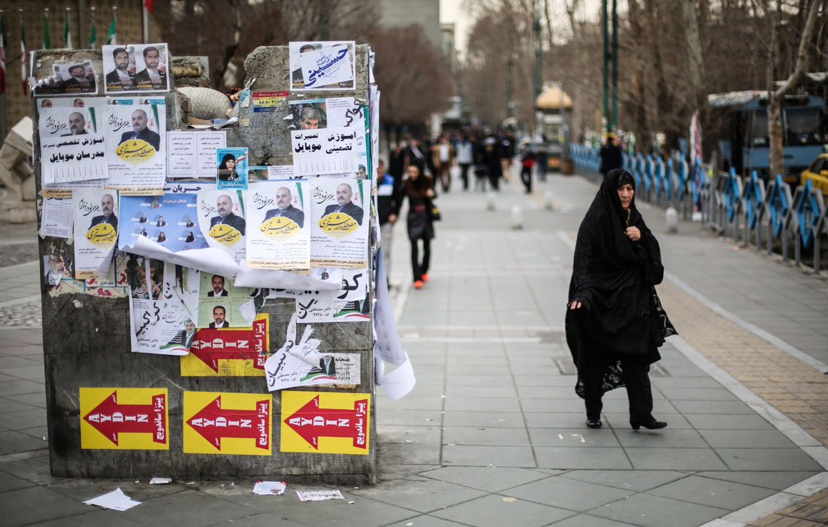 تظهر الصورة مشهداً في منطقة حضرية، حيث تُظهر مجموعة من الملصقات الإعلانية المعلقة على جدار، تتضمن صوراً وعبارات مكتوبة. في المقدمة، توجد امرأة ترتدي حجاباً أسود، تسير في اتجاه معين. الطريق مزدحم بعض الشيء، مع وجود أشخاص آخرين في الخلفية ووسائل النقل. يبدو أن الجو يميل إلى البرودة، ويمكن رؤية الأشجار والبنية التحتية الحضرية.