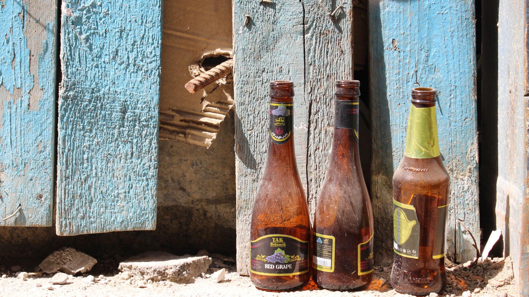 The image features three glass bottles positioned in front of a weathered blue wooden wall. The bottles appear dusty and have some remnants of liquid inside them. The wall shows signs of decay, with peeling paint and a small gap revealing damaged wood behind it. The ground is uneven, with dirt and debris scattered around, adding to the overall rustic and neglected feel of the scene.
