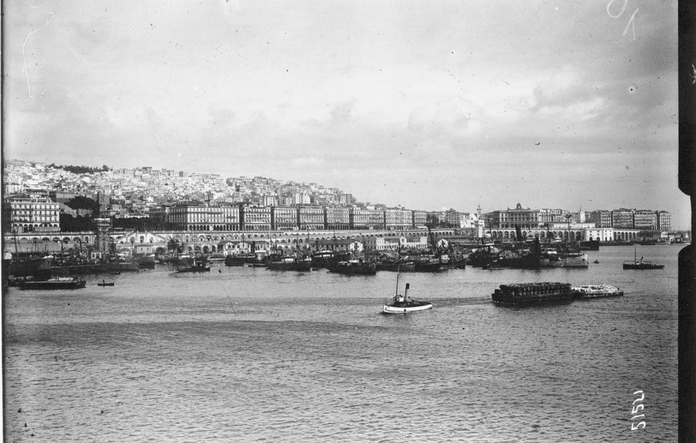 L'image représente un port où l'on peut voir plusieurs bateaux amarrés. Au fond, une ville s'étend, avec des bâtiments alignés sur la côte. Le ciel est nuageux, et l'eau calme reflète la scène. Les collines derrière la ville sont habitées, ajoutant à la densité urbaine de l'endroit. L'ambiance générale semble nostalgique, probablement en raison du style photographique ancien.