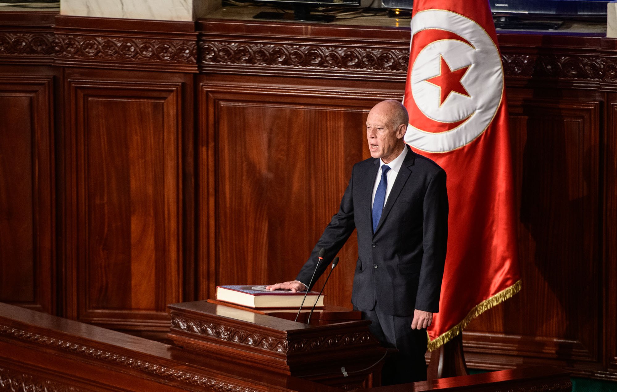 The image depicts a formal setting, likely in a government assembly or parliament. A man, dressed in a dark suit and tie, stands at a podium with a large book placed on it. Behind him, there is a prominent flag featuring a red background with a white circle and a red crescent and star, indicating it is Tunisia's national flag. The environment appears ornate, with wooden paneling and an overall serious ambiance, suggesting that he is addressing a legislative body or delivering an important speech.