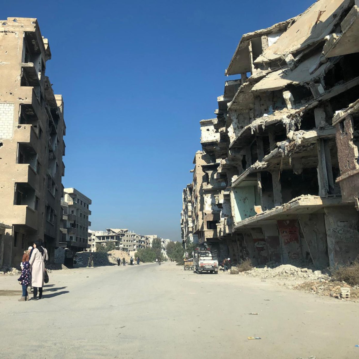 تصویر یک خیابان خالی را نشان می‌دهد که در دو طرف آن ساختمان‌های مخروبه و آسیب‌دیده قرار دارند. دیوارهای این ساختمان‌ها پر از سوراخ‌ها و آسیب‌های ناشی از جنگ یا تخریب هستند. در انتهای خیابان، چند ساختمان دیگر نیز مشاهده می‌شود. آسمان آبی و صاف است و افراد کمی در خیابان در حال تردد هستند. دو نفر در سمت چپ تصویر به نظر می‌رسد در حال صحبت کردن هستند و یک کودک نیز با آن‌هاست. فضای کلی تصویر تنهایی و تخریب را القا می‌کند.