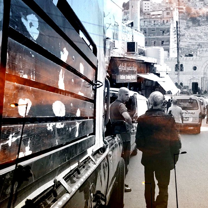 تبدو الصورة عبارة عن مشهد حضري يظهر شارعًا مزدحمًا حيث يسير أحد الأشخاص بعصا مساعدة. تظهر في الخلفية مباني متعددة الأشكال، وبعض السيارات تسير في الشارع. الأجواء تعكس حركة الحياة اليومية، ويبدو اللون الرمادي والأسود مسيطرًا مع لمسات من الألوان الدافئة، مما يضفي شعورًا بالحنين أو الذكريات.