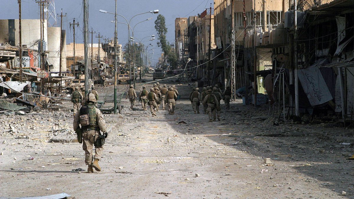 تظهر الصورة مشهدًا لمدينة مدمرة، حيث تسير مجموعة من الجنود في شارعٍ واسع. الشوارع تبدو خالية وتظهر فيها آثار الدمار، مثل المباني المتضررة والأدوات المتروكة. السماء تبدو غائمة، مما يضفي على المشهد طابعًا قاتمًا. الجنود يرتدون زيًا عسكريًا ويحملون معداتهم، مما يشير إلى أنهم في مهمة عسكرية.