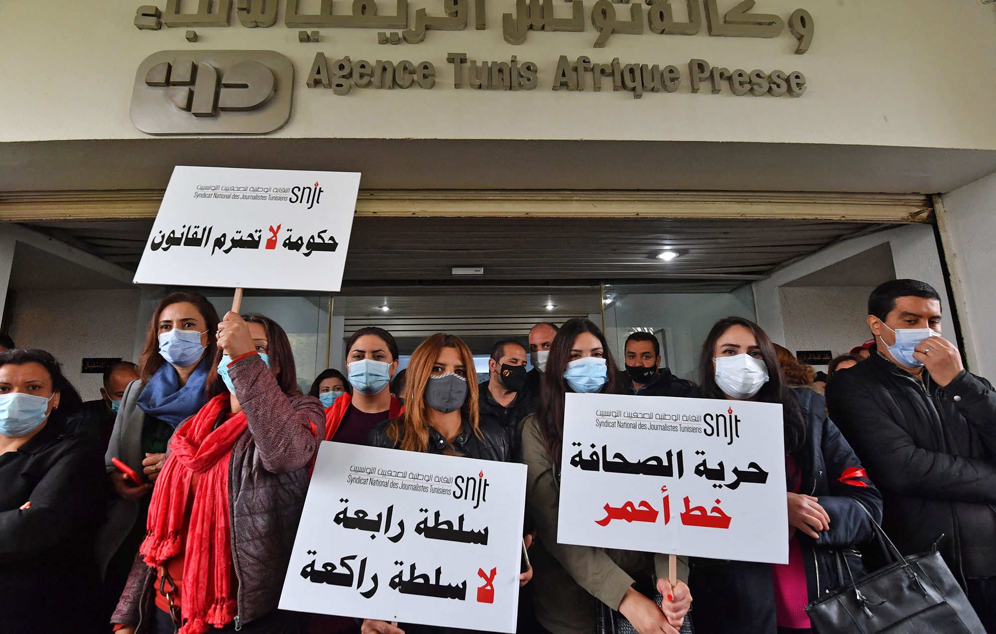 L'image montre un groupe de femmes et d'hommes rassemblés devant un bâtiment, portant des pancartes. Les pancartes expriment des messages liés à la liberté de la presse et critiquent le gouvernement. Les manifestants portent des masques, probablement en raison de préoccupations sanitaires. En arrière-plan, on peut voir le logo d'une agence de presse. L'atmosphère semble déterminée, avec un engagement visible pour des causes liées à la liberté d'expression.