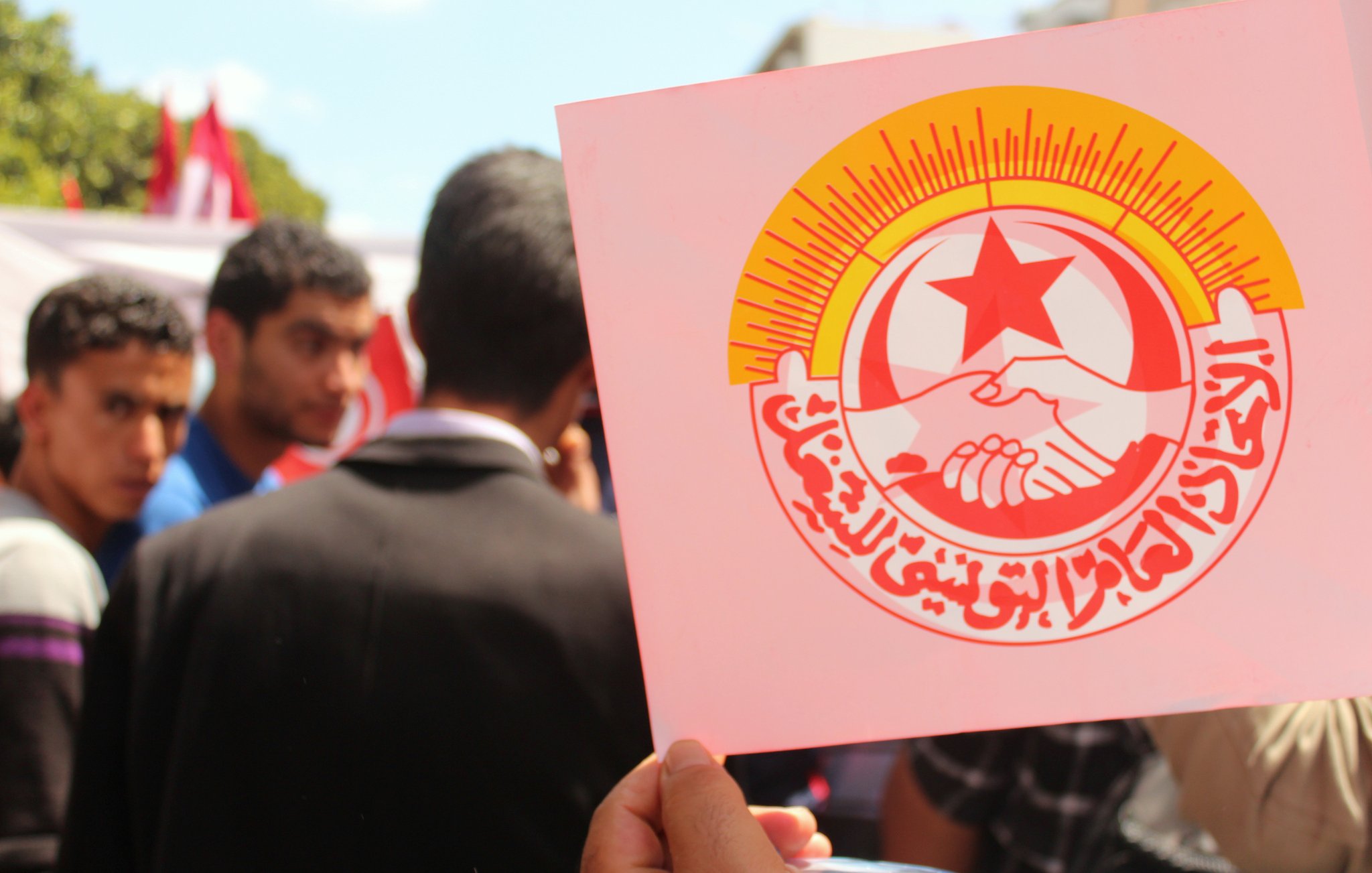 L'image montre une manifestation ou un rassemblement où une personne tient une pancarte avec un logo. Ce logo représente probablement une organisation syndicale ou politique, avec des éléments symboliques comme des mains qui se serrent et une étoile rouge. En arrière-plan, on peut apercevoir des gens qui semblent participer à l'événement, ainsi que des drapeaux. L'atmosphère semble être celle de solidarité et de mobilisation.