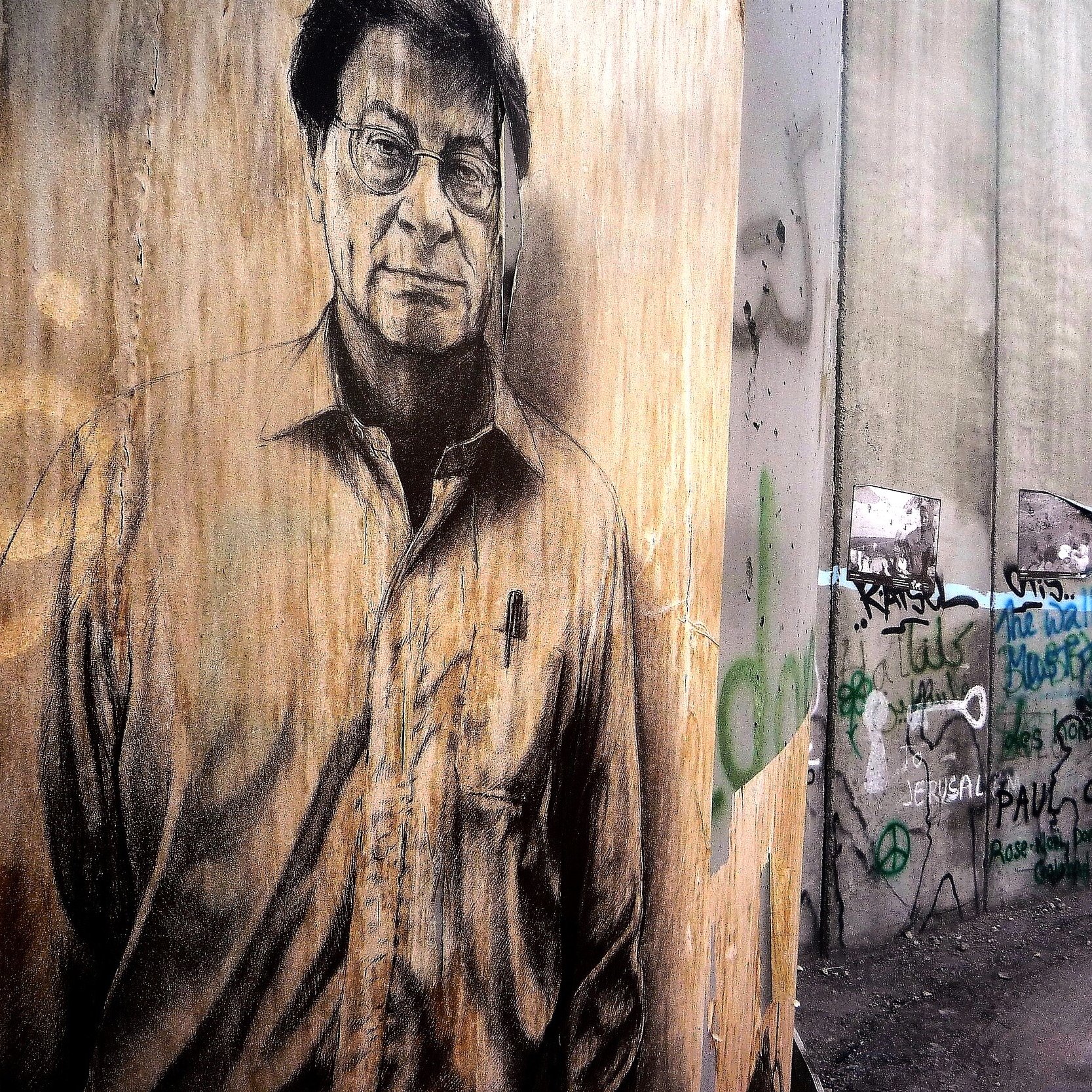 L'immagine rappresenta un murale su un muro di cemento, caratterizzato da un ritratto di un uomo con occhiali e un'espressione seria. Il muro è grezzo e presenta anche scritte e graffiti, suggerendo un contesto urbano e di protesta. Il contrasto tra il ritratto e il resto del muro evidenzia un messaggio artistico e sociale. La scena evoca un senso di riflessione e di critica.