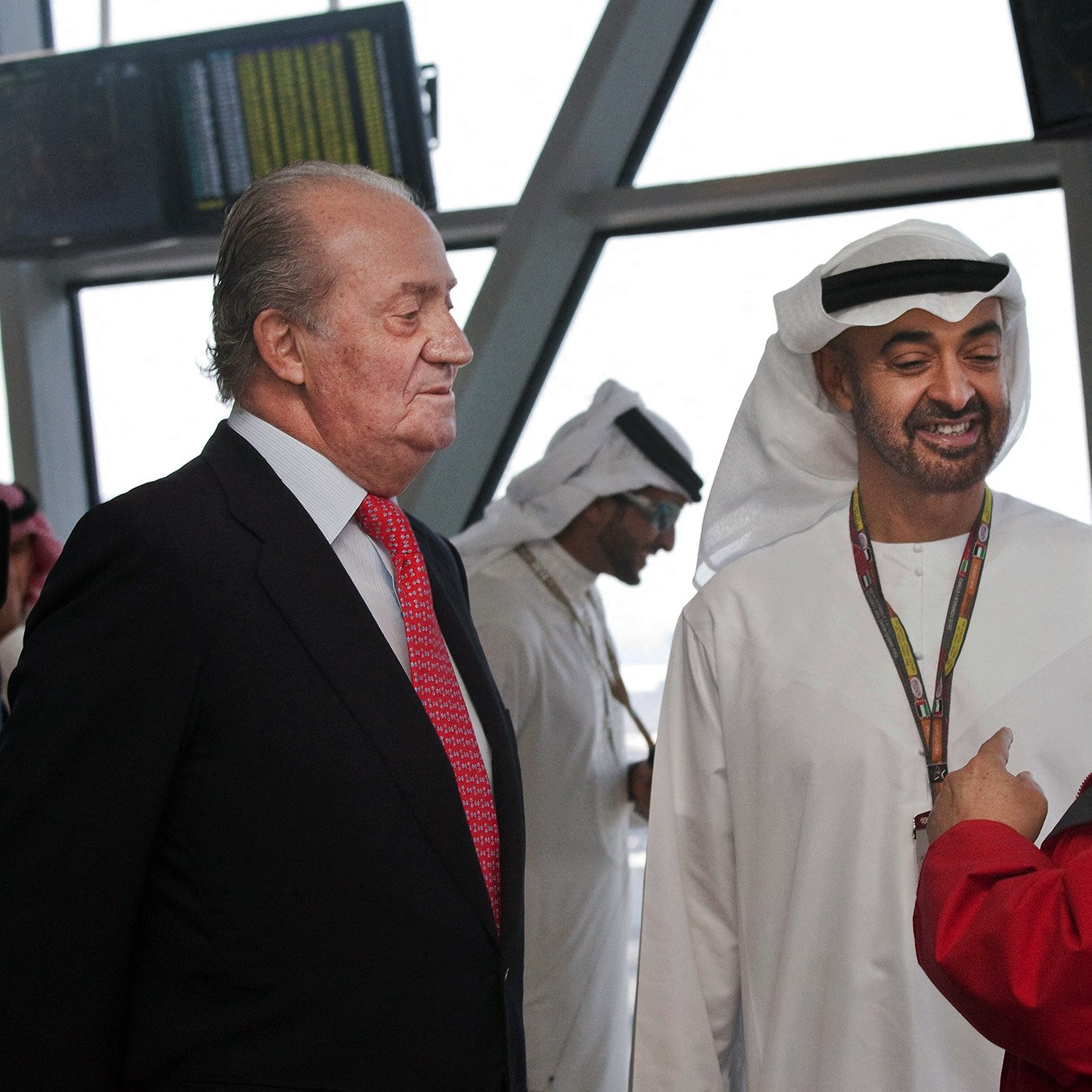 En la imagen se pueden ver a tres hombres conversando. Dos de ellos visten trajes tradicionales árabes, mientras que el tercero lleva un traje negro con una camisa blanca y una corbata roja. El entorno parece ser un aeropuerto o un lugar moderno. Los hombres están interactuando de manera amigable y hay pantallas en el fondo, lo que sugiere un ambiente ocupado y profesional.