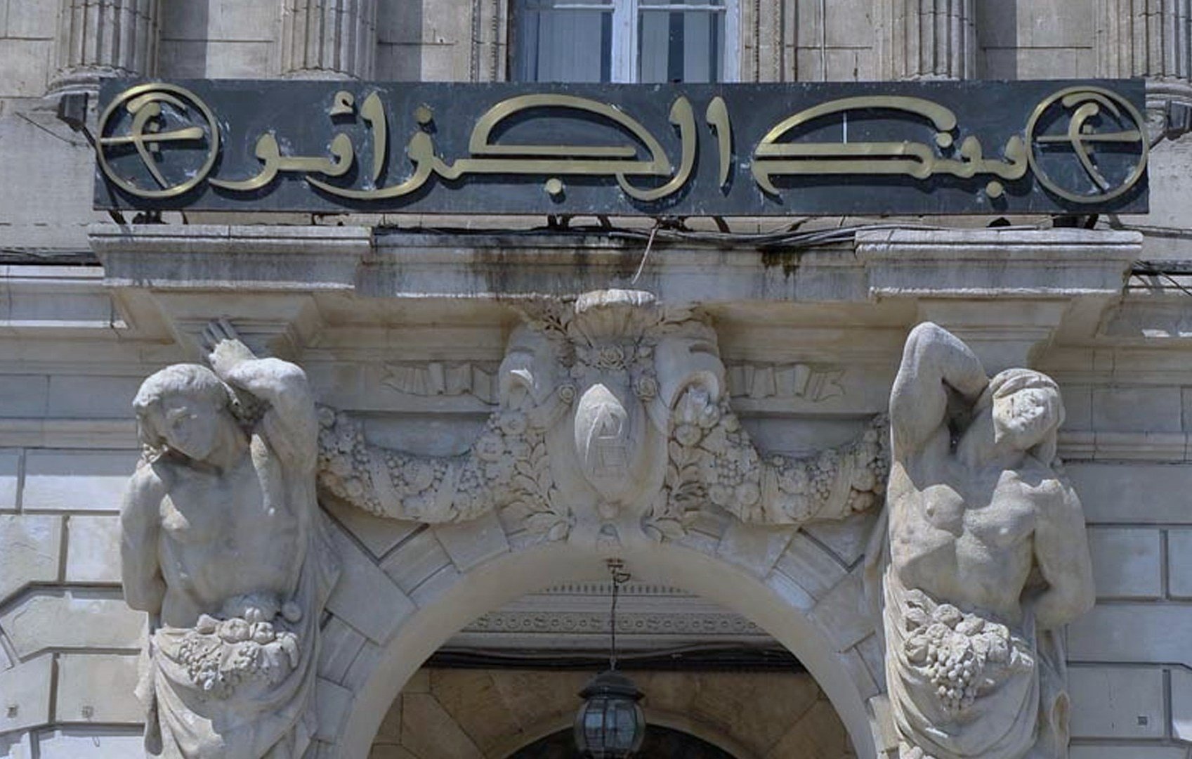 The image features a historical bank building with an ornate entrance. Above the doorway, there is a sign in Arabic that reads "بنك الجزائر" which translates to "Bank of Algeria." The architecture includes decorative elements, such as sculptures of two male figures on either side of the entrance. These figures appear to be holding or surrounded by garlands or fruit, adding to the classical decorative style of the building. The overall design is elaborate, reflecting a historical architectural style.