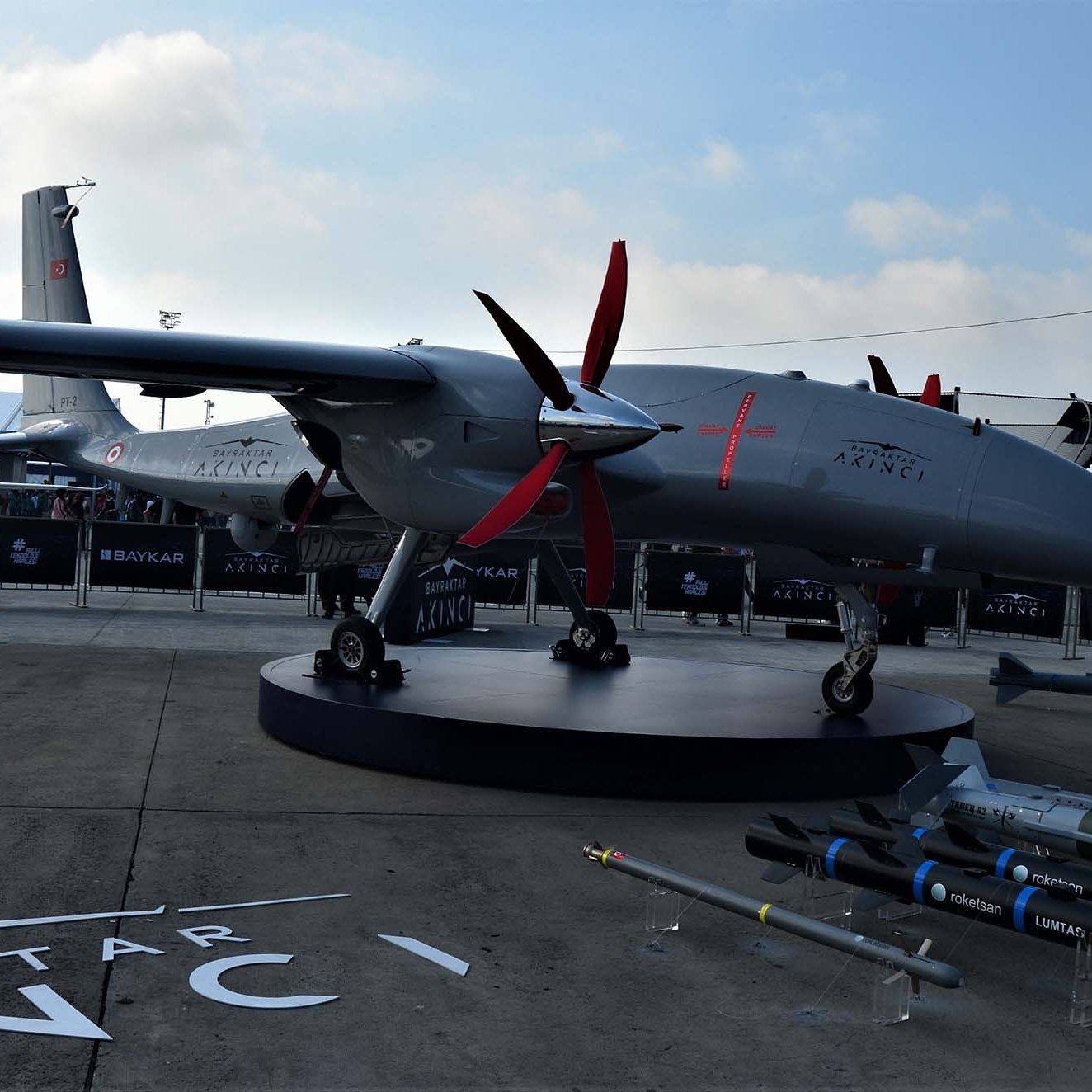 این تصویر نمایش یک پهپاد نظامی به نام "بایراکتار" است که در یک نمایشگاه هوایی یا نظامی به نمایش گذاشته شده است. پهپاد دارای طراحی مدرن و بدنه‌ای خاکستری رنگ است. پروانه‌های قرمزرنگ در قسمت جلو، زیبایی و قدرت آن را نشان می‌دهند. همچنین در کنار پهپاد، موشک‌ها و سلاح‌های مختلفی قرار دارند که به قابلیت‌های نظامی این پهپاد اشاره دارند. در پس‌زمینه، چادرها و سازه‌های نمایشگاهی قابل مشاهده هستند. آسمان کمی ابری به نظر می‌رسد.
