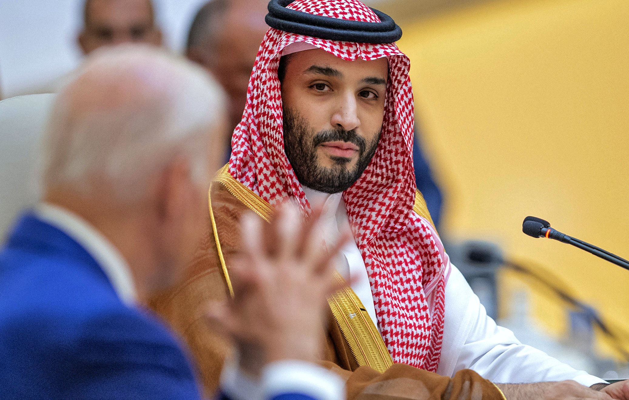 L'image montre un homme portant une tenue traditionnelle saoudienne, comprenant un thobe et un ghutrah (un couvre-chef en tissu). Il est assis à une table lors d'une réunion, fixant le spectateur avec un regard sérieux. À l'arrière-plan, un autre homme, dont le visage est partiellement visible, semble être en train de parler ou de poser une question. L'environnement suggère un cadre formel et officiel, comme une conférence ou un sommet.