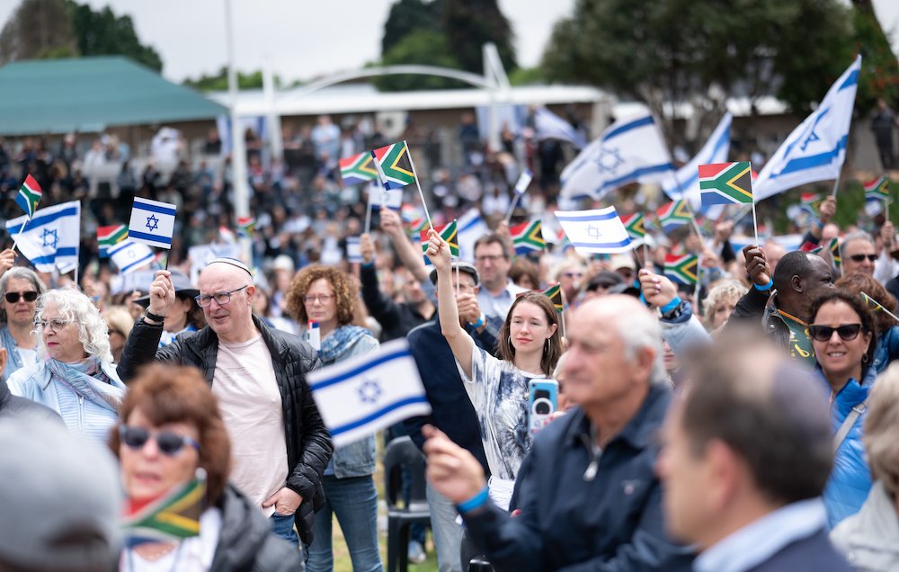 در این تصویر، گروهی از مردم در یک مراسم یا تجمع بزرگ دیده می‌شوند. آن‌ها در حال تکان دادن پرچم‌های اسرائیل و آفریقای جنوبی هستند. جمعیت شامل مردان و زنانی از تنوع سنی مختلف است و برخی از آن‌ها در حال لبخند زدن و خوشحالی دیده می‌شوند. فضای عمومی کمی ابری به نظر می‌رسد و تعداد زیادی کرسی در پس‌زمینه وجود دارد.
