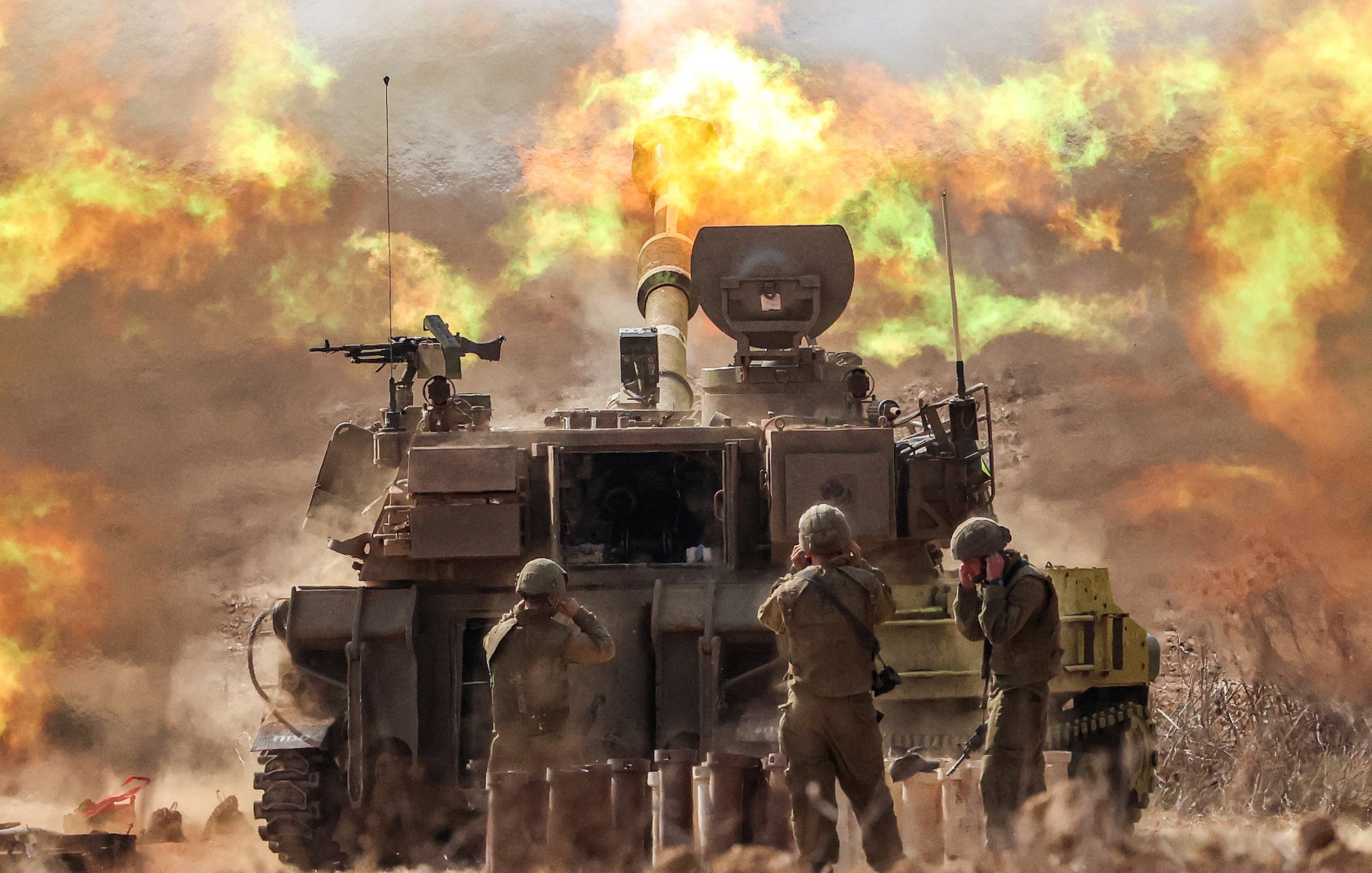 L'image montre un char de combat en action, probablement en train de tirer. Des soldats en uniforme sont présents près du char, occupant des positions de tir ou se préparant à l'action. En arrière-plan, des explosions et des flammes se produisent, créant une atmosphère intense et chaotique. La scène évoque un environnement militaire, avec une ambiance de conflit et de mouvement.