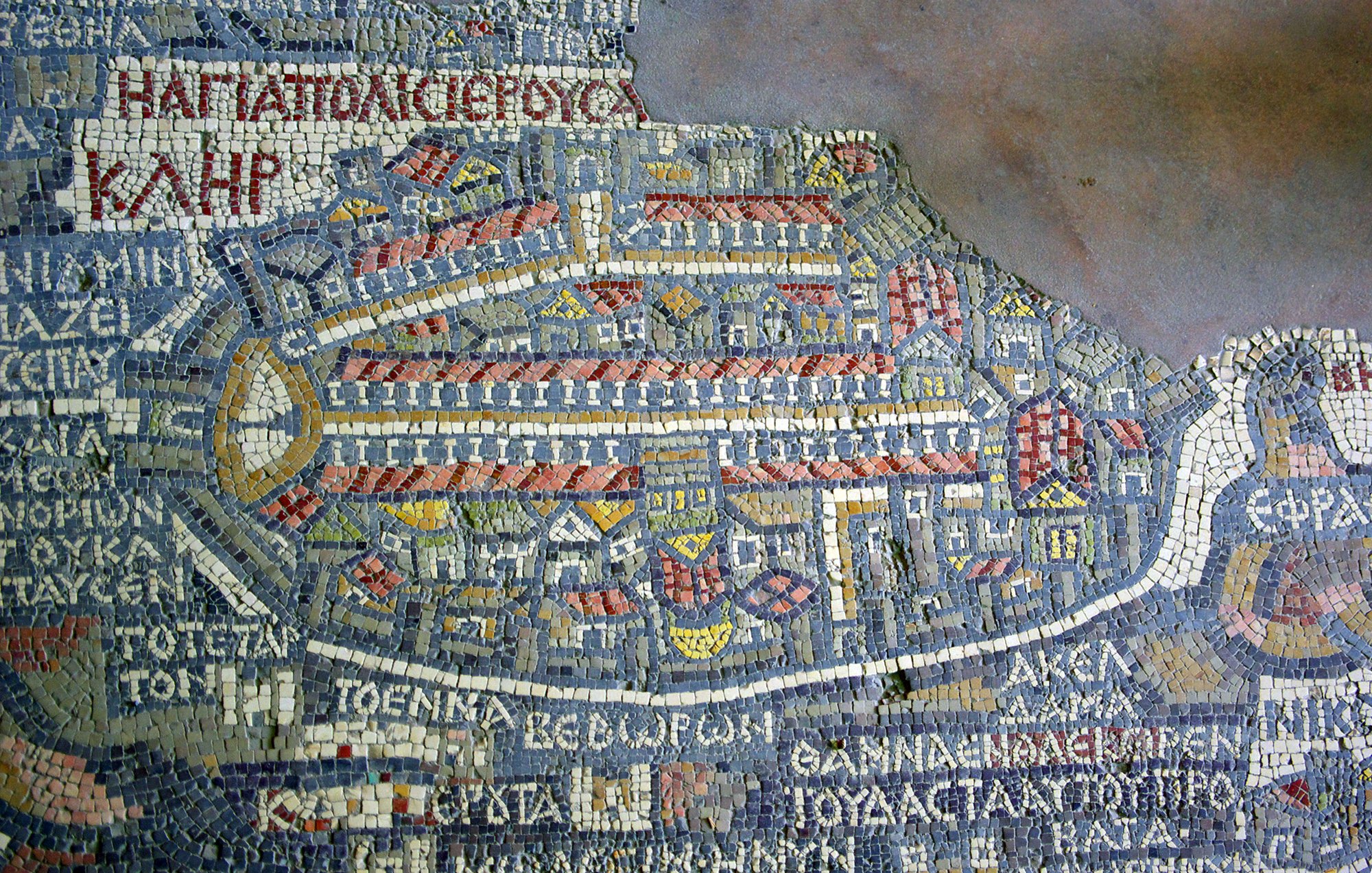 The image depicts a mosaic map, featuring intricate tile work that illustrates an ancient city layout. The map is composed of various colored tiles, forming buildings, streets, and outlines of the architecture within the city. It includes labeled regions or areas, indicating places of significance in the city, which might be historical or prominent during the time it represents. The overall design reflects a detailed representation of urban planning, showcasing the artistry and craftsmanship of the era.
