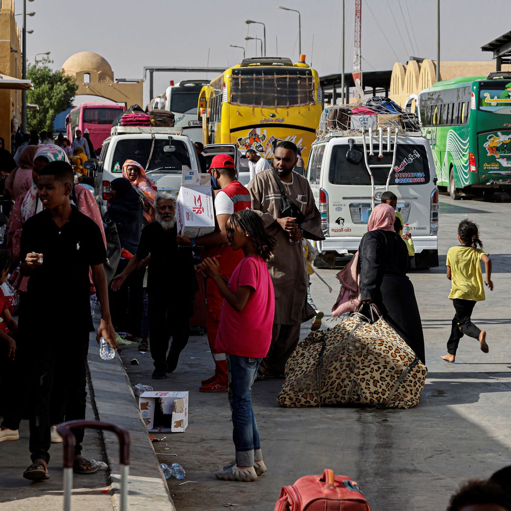 La imagen muestra una estación de autobuses donde hay un gran movimiento de personas. Se puede ver a varias familias y grupos de personas caminando entre los autobuses, algunos con maletas y bultos. Hay un ambiente de actividad, con adultos y niños, algunos consumiendo bebidas y otros llevando objetos. En el fondo, se aprecian varios autobuses de diferentes colores, indicando que podría ser un lugar de transporte importante. La escena refleja un momento de desplazamiento y espera.