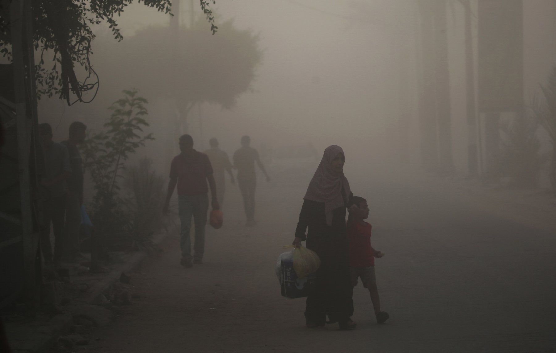 L'image montre une scène urbaine plongée dans une atmosphère brumeuse ou poussiéreuse. Dans le premier plan, une femme portant un hijab tient la main d'un jeune garçon. Ils semblent marcher ensemble le long de la rue. En arrière-plan, d'autres silhouettes de personnes sont visibles, floues par la brume, tandis que des arbres et des structures urbaines apparaissent à peine à travers la poussière. L'ambiance semble calme mais chargée d'un certain mystère à cause de la visibilité réduite.