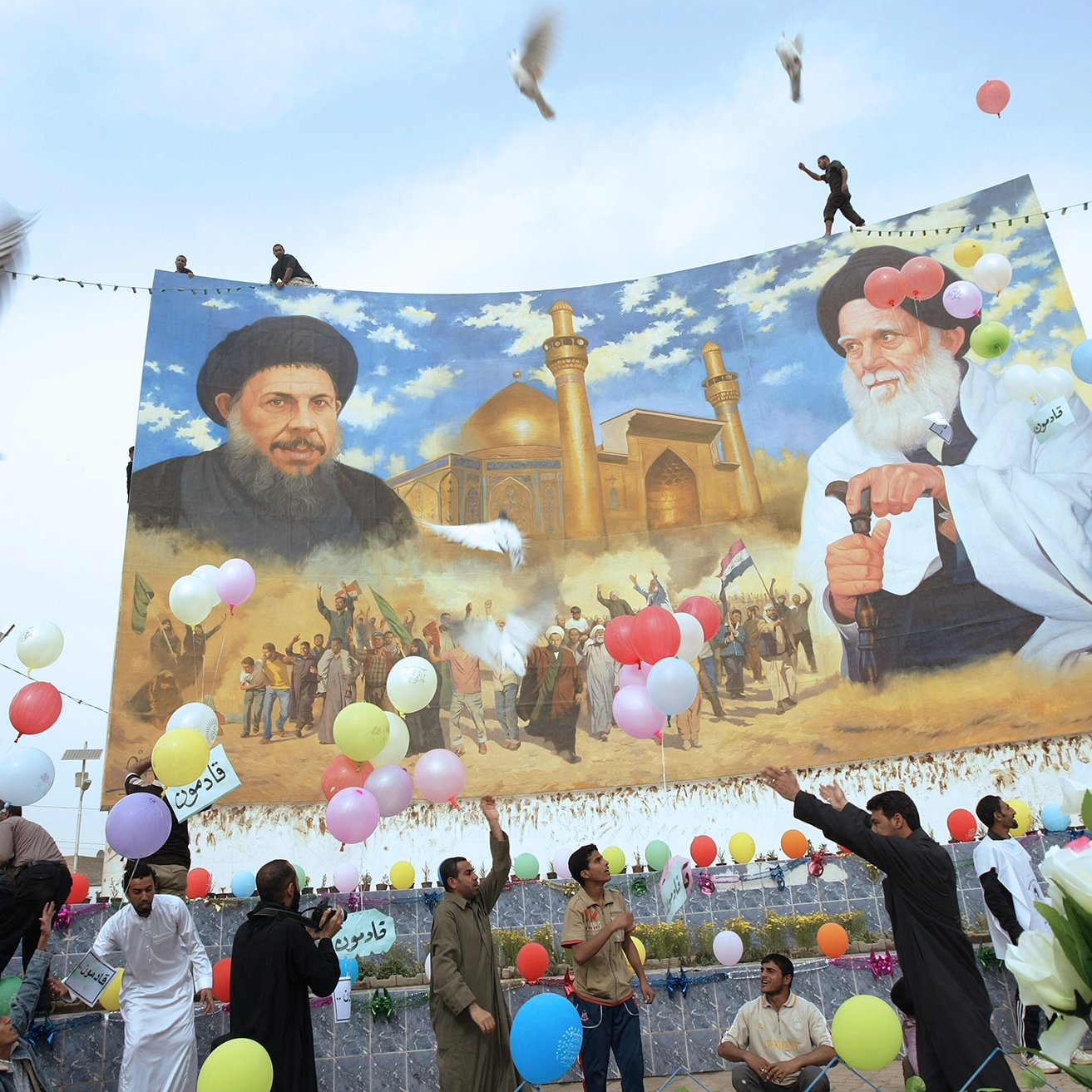 L'image montre une scène festive en extérieur avec un grand panneau représentant deux figures, probablement des leaders ou des personnalités importantes. Au-dessous de ce panneau, des gens célèbrent, tenant des ballons colorés et des fleurs. Certaines personnes semblent danser ou exprimer leur joie. En arrière-plan, il y a un décor architectural qui semble être un bâtiment religieux, avec des minarets visibles. Des colombes volent également dans le ciel, ajoutant une dimension symbolique à l'événement.