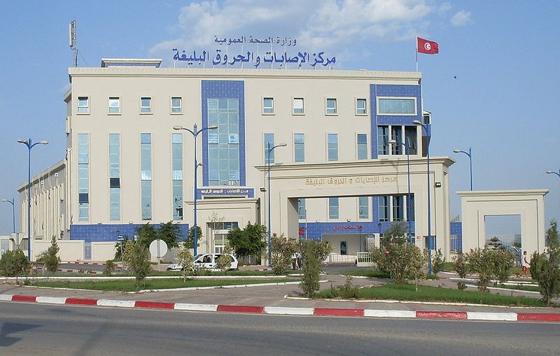 L'image montre un bâtiment moderne, probablement un centre de santé ou une institution gouvernementale. La façade est de couleur claire avec des éléments architecturaux en bleu. On y voit des fenêtres de différentes tailles et une grande enseigne en arabe et en français. Un drapeau tunisien flotte sur le toit, ce qui indique que le bâtiment est situé en Tunisie. L'environnement semble soigné, avec un espace urbain bien aménagé autour du bâtiment.
