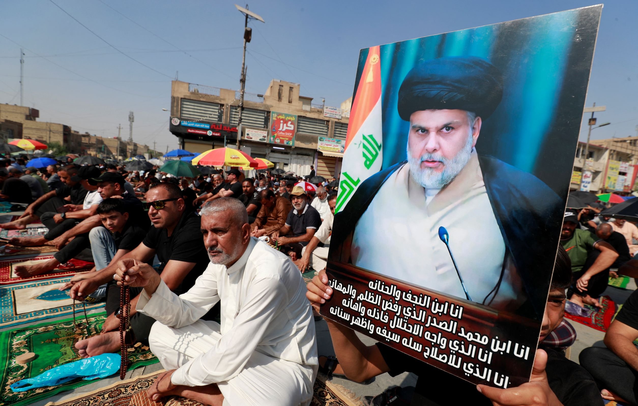 The image depicts a large gathering of people, seated on mats in an outdoor setting, likely in a city square. Many attendees are dressed in black, which is often associated with mourning or commemorative events. One man stands out, holding a prominent poster featuring a portrait of an individual wearing traditional attire, along with a flag in the background. The atmosphere suggests a significant religious or political event, characterized by unity and solemnity among the participants.