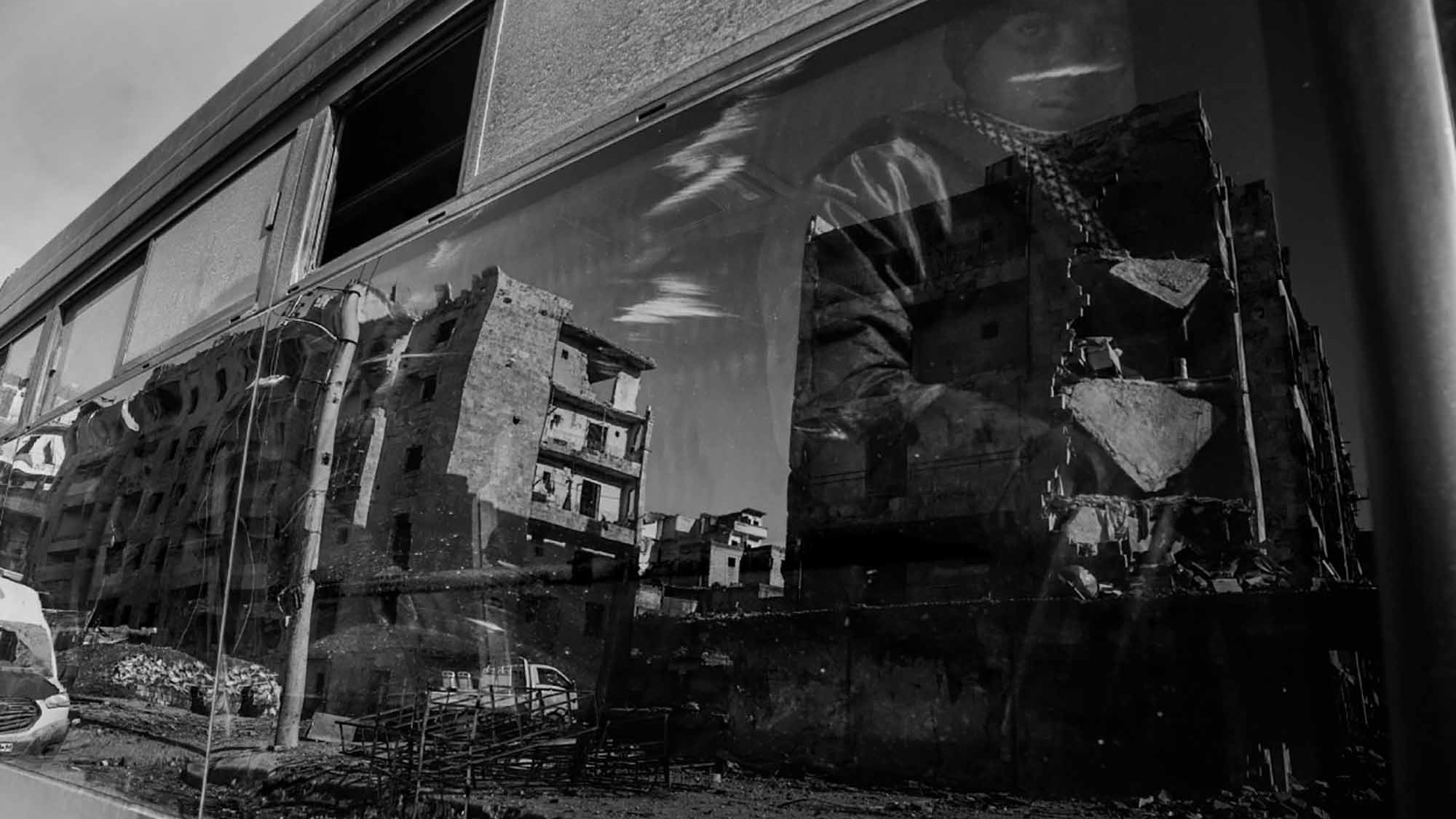 L'image présente une scène en noir et blanc, où un bus est au premier plan. On peut voir le reflet d'un homme, probablement en train de porter un vêtement sombre et une écharpe, à côté d'une architecture en ruine. À travers la vitre du bus, des bâtiments dégradés sont visibles, indiquant des signes de destruction. L'atmosphère semble sombre et tragique, évoquant des thèmes de dévastation et de résilience.