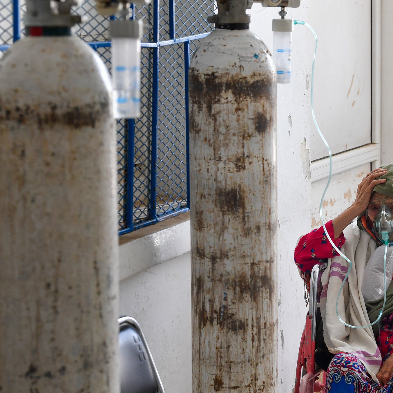 L'immagine mostra una donna anziana seduta su una sedia a rotelle in un ambiente ospedaliero. Indossa una maschera per l'ossigeno e sembra preoccupata, con una mano sulla fronte. Dietro di lei ci sono bombole di ossigeno e un ambiente che appare abbastanza semplice e clinico. L'atmosfera trasmette una sensazione di vulnerabilità e dolore, evidenziando le difficoltà legate alla salute.