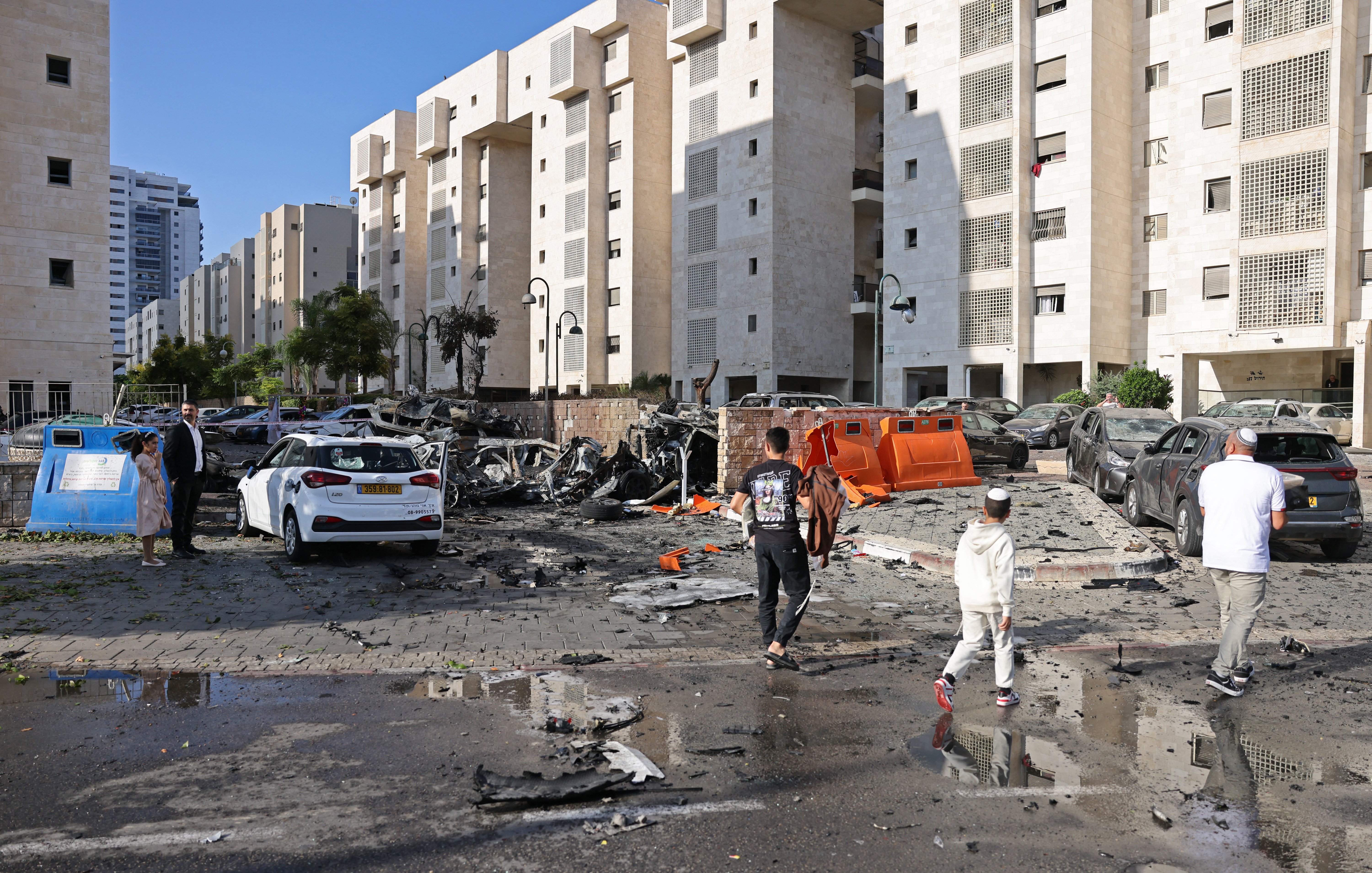L'immagine mostra una scena di distruzione in un'area urbana. Si possono vedere edifici residenziali con facciate chiare, circondati da macerie e detriti. Alcune auto sono danneggiate, con pezzi sparsi sul terreno. Ci sono persone che camminano nella strada, apparentemente mentre osservano il danno. Il cielo è sereno, e ci sono anche pozzanghere d'acqua, suggesting che recente pioggia potrebbe essere rientrata nell'area. La scena trasmette un senso di caos e di recente violenza o attacco.