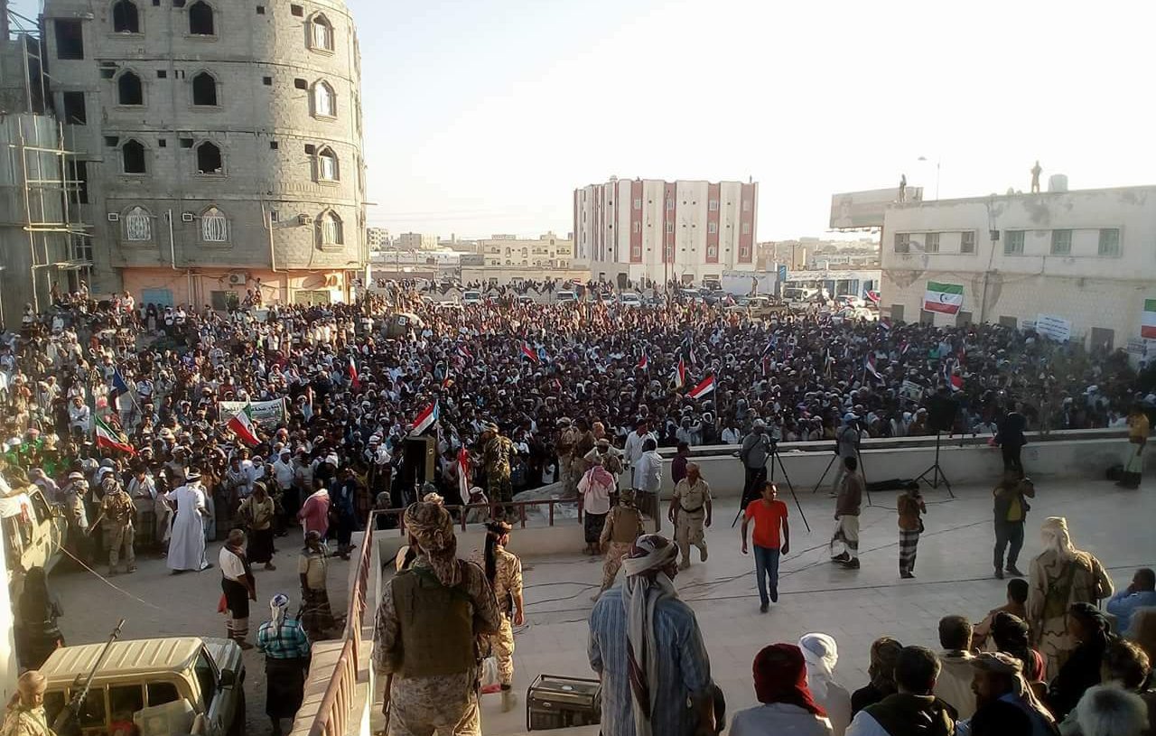 الصورة تظهر تجمعًا حاشدًا من الناس في مكان عام، حيث يبدو أنهم يشاركون في فعالية أو تظاهرة. المجموعة كبيرة وتحتوي على عدد من الأعلام، مما يشير إلى أن هناك موضوعًا معينًا يجمعهم. المباني المحيطة توحي بأن المكان في منطقة حضرية، كما أن بعض الأشخاص يجلسون على الدرج بينما يتواجد آخرون في المقدمة. تبرز الأجواء الحماسية في الحشد.
